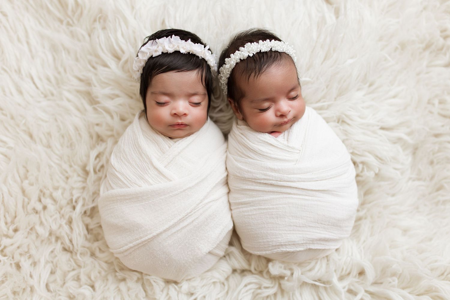 Twin newborns wrapped in white swaddles with headbands lay together on fluffy white fur.