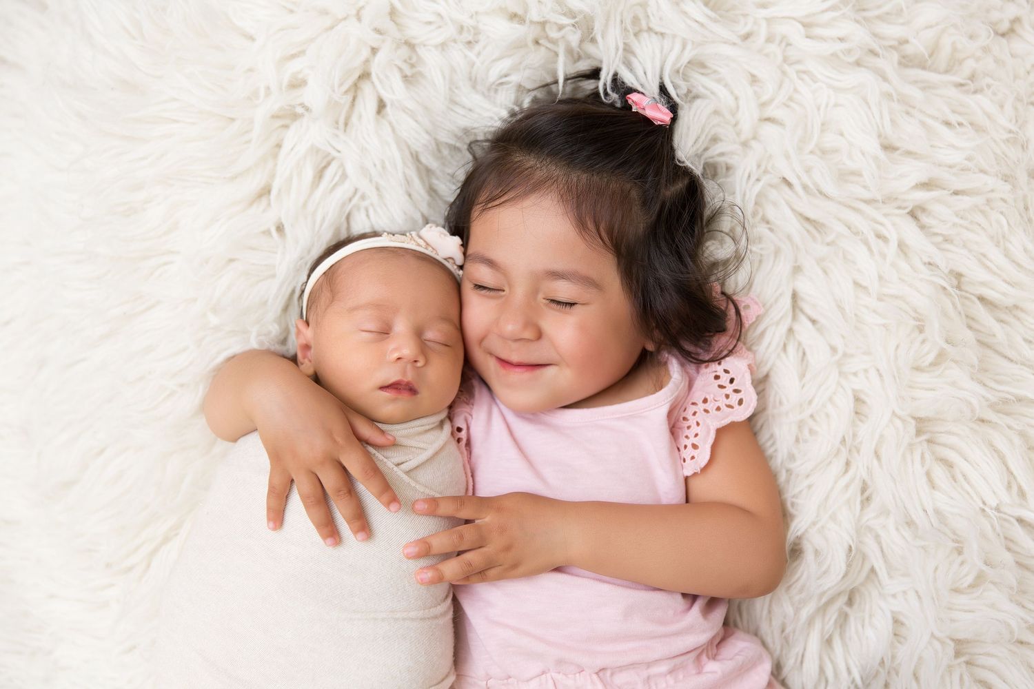 Sweet sibling moment on white fluffy rug with sleeping newborn and sister in pink.