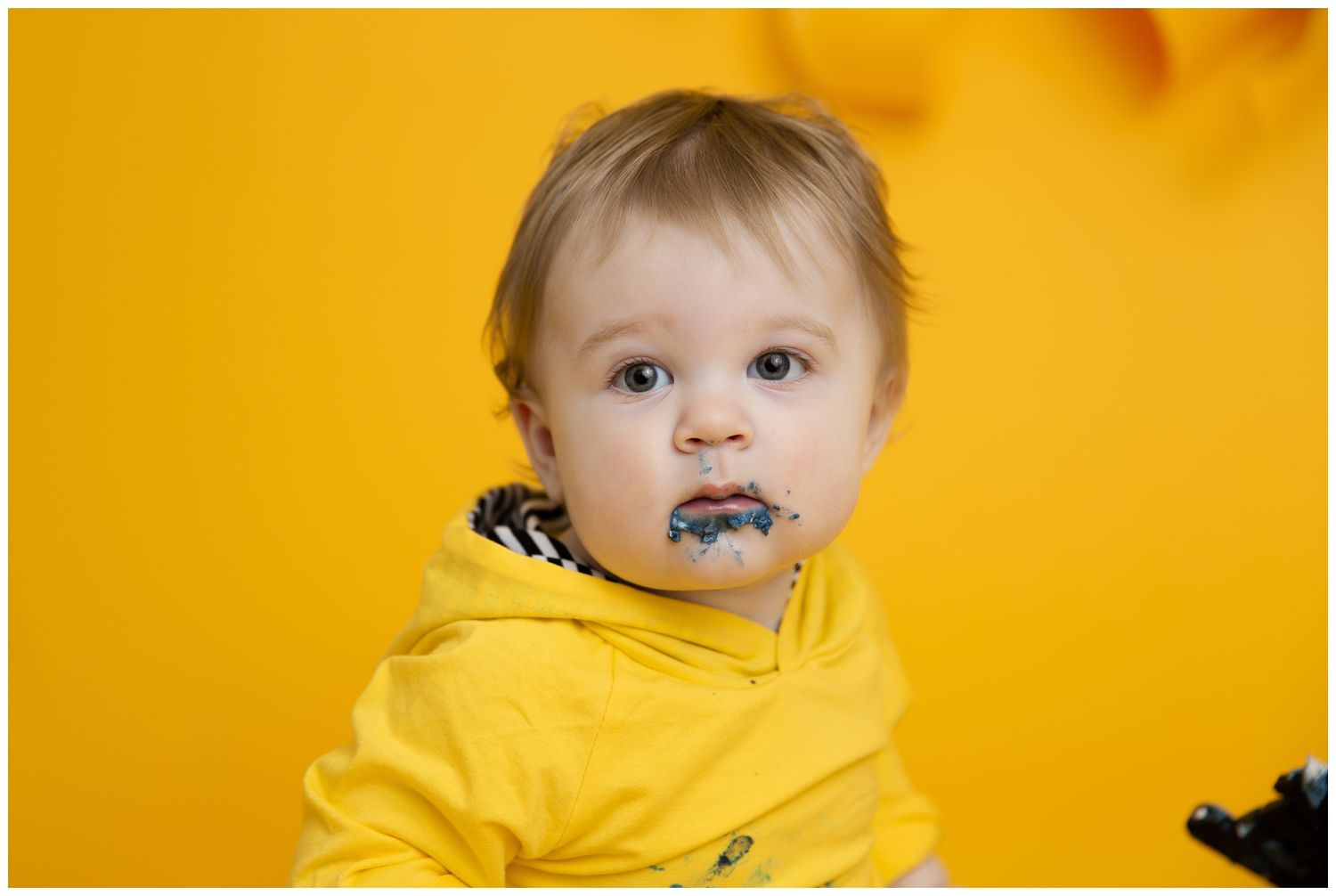 A small child in a bright yellow hoodie against an orange background with a blue pacifier in their mouth.