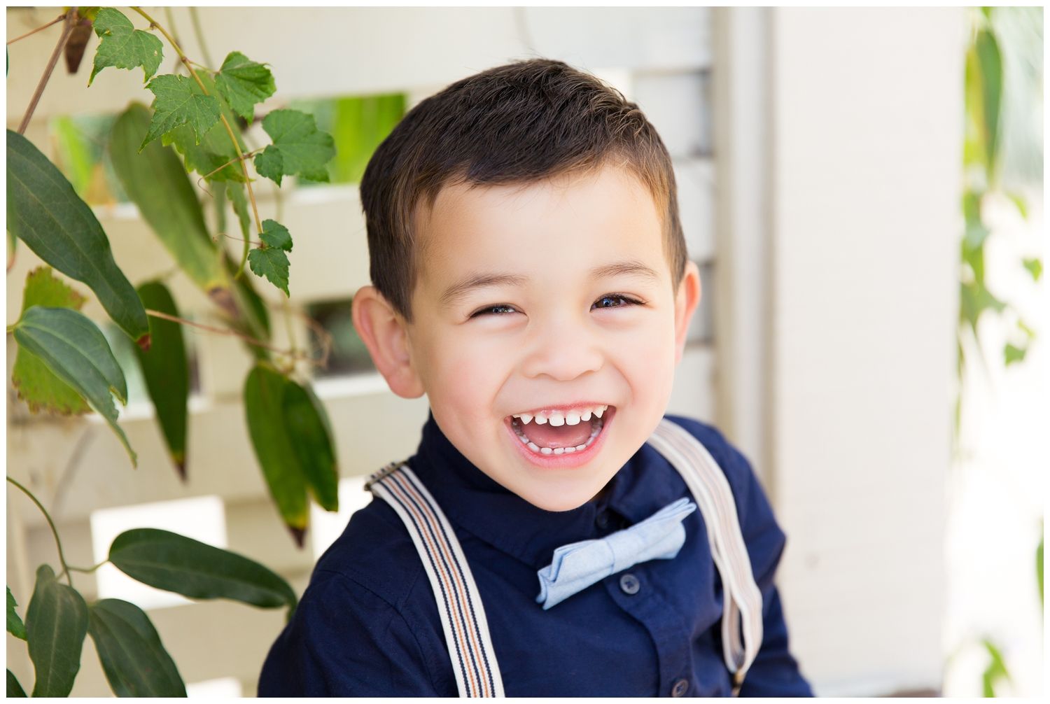 A young child wearing suspenders and a white shirt smiles brightly while sitting near a window with plants.