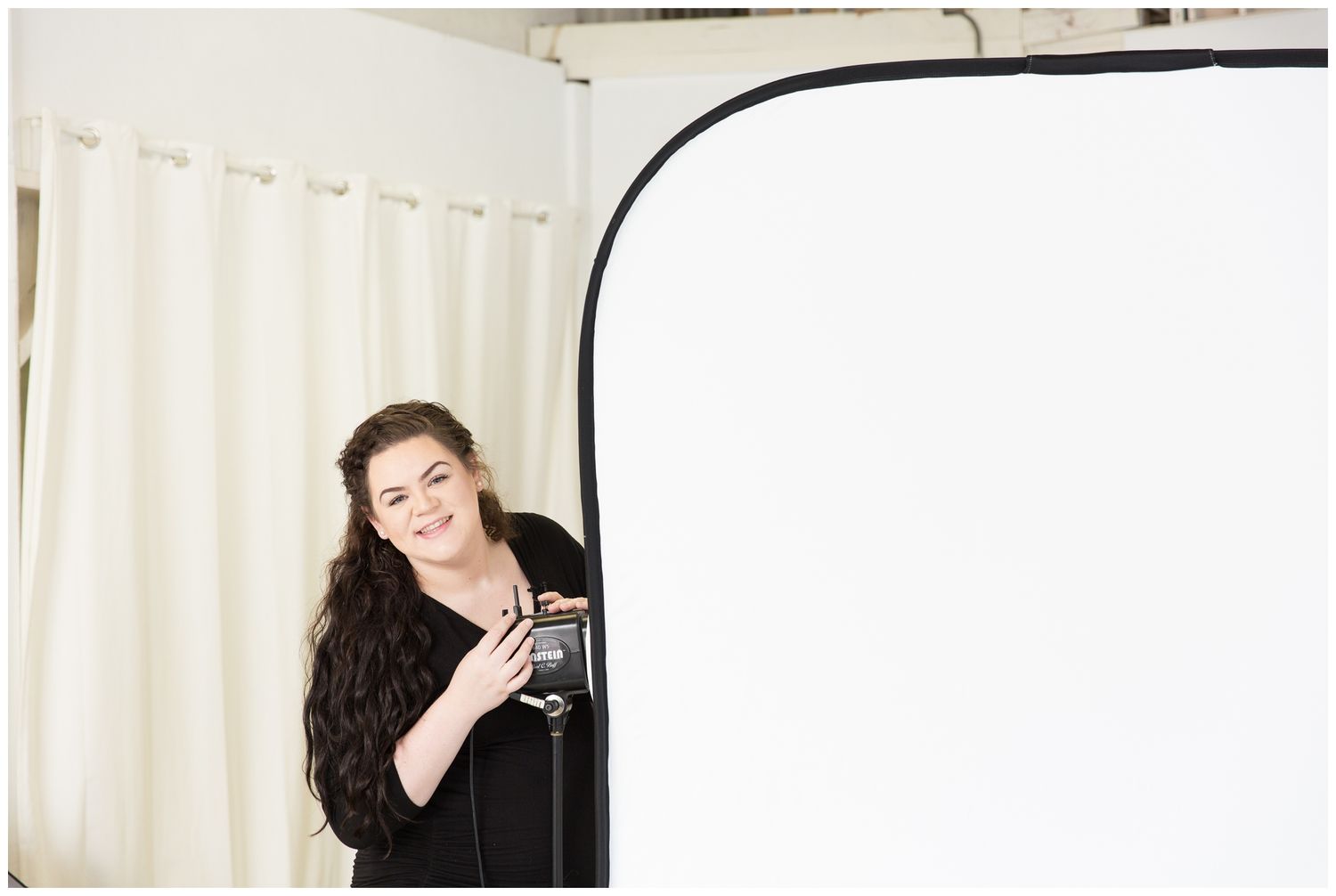 A photographer peeks out from behind a white backdrop while holding a vintage camera.