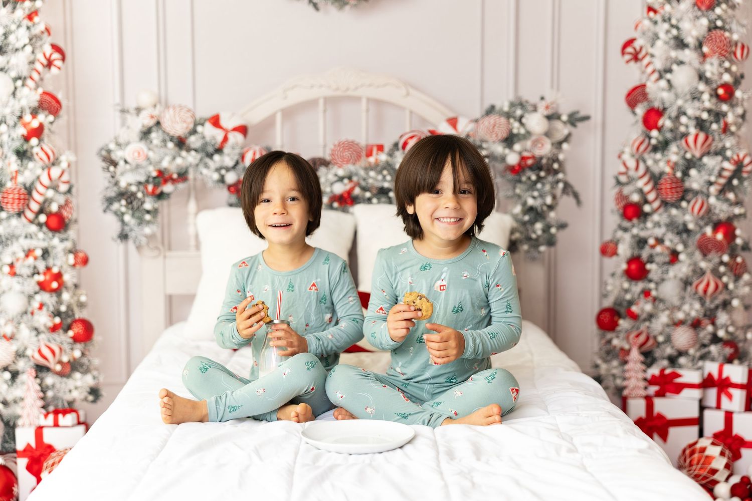Two children in matching blue pajamas sitting on bed with Christmas decorations.