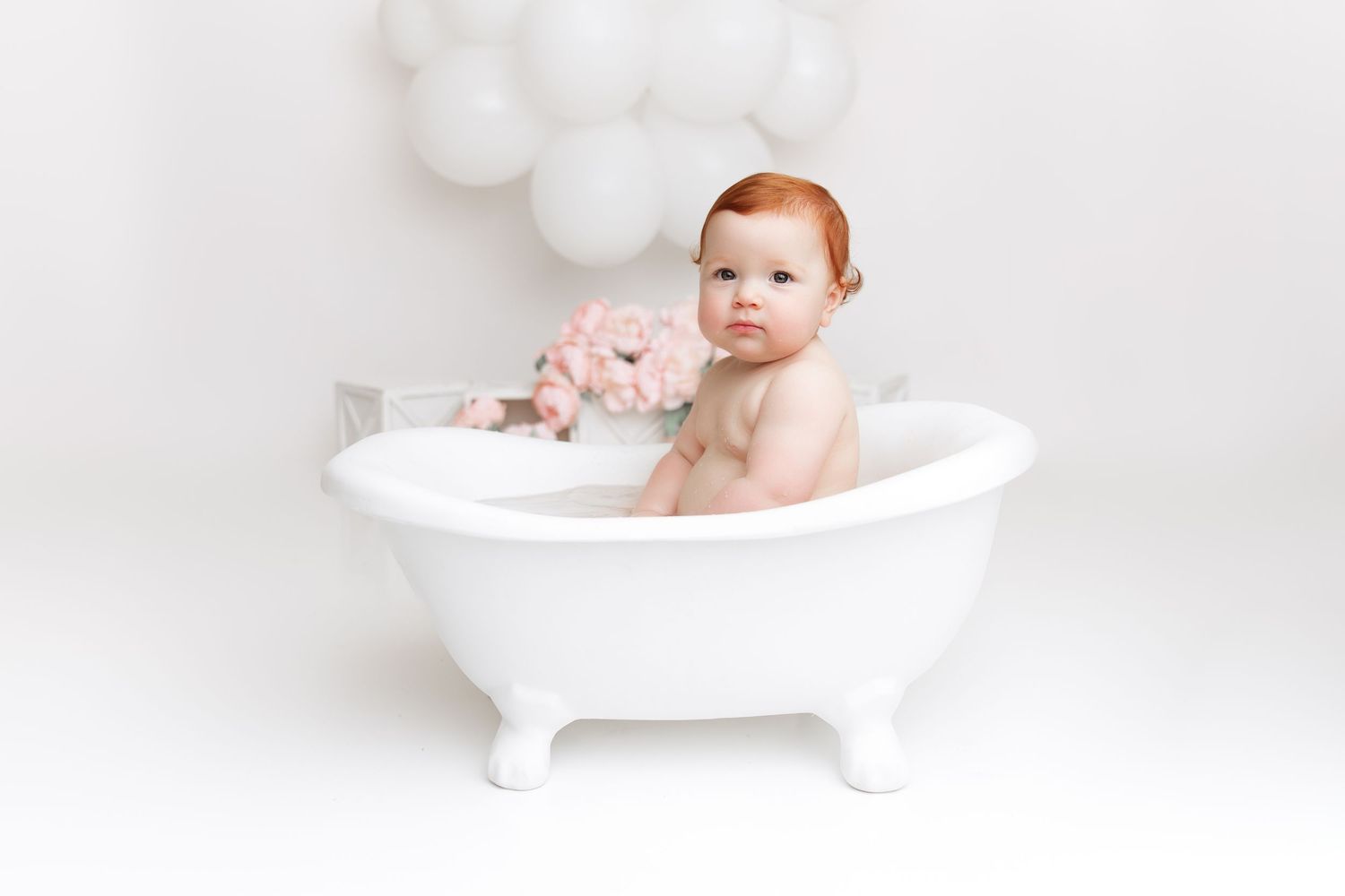Baby sitting in white clawfoot bathtub with white balloon decorations in background.
