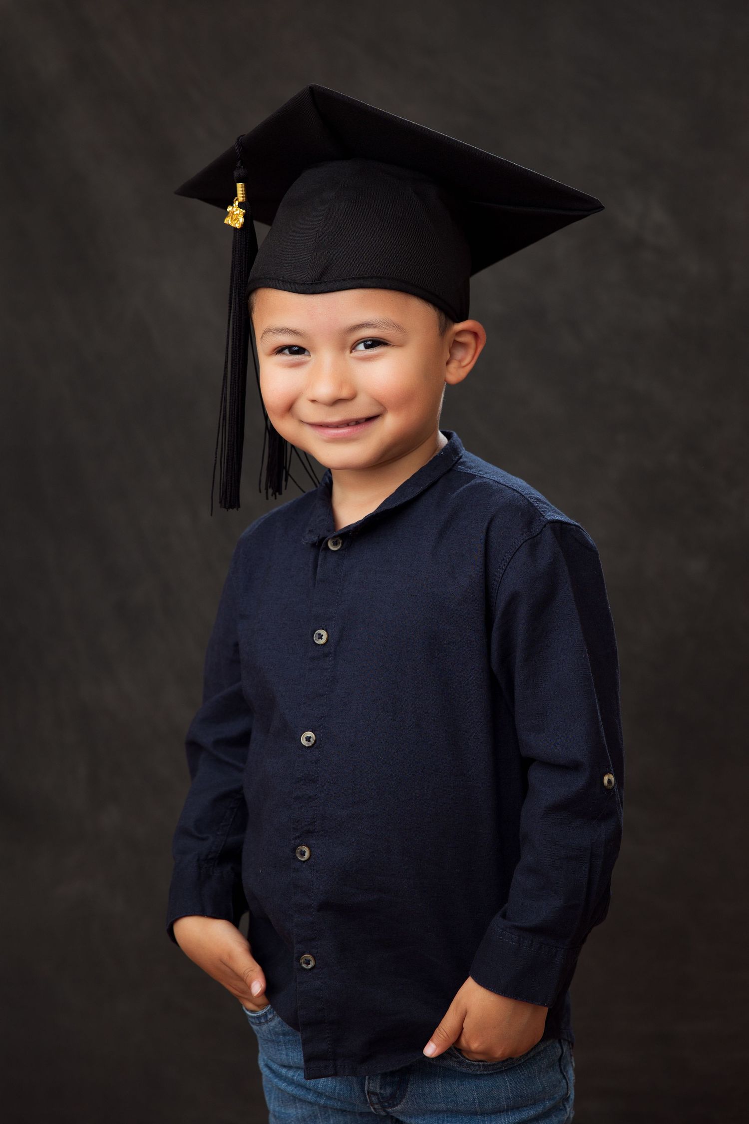 Young graduate wearing a mortarboard cap, navy button-up shirt and jeans poses for a portrait against dark background.