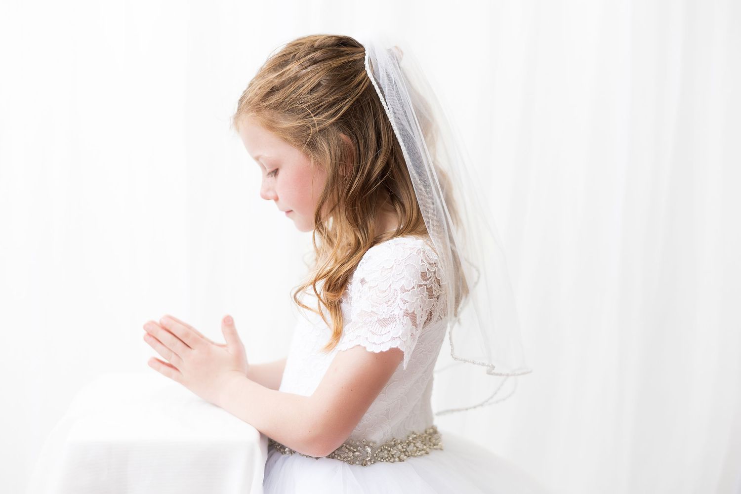 A young figure in a white lace communion dress with veil stands in prayer against a bright background.