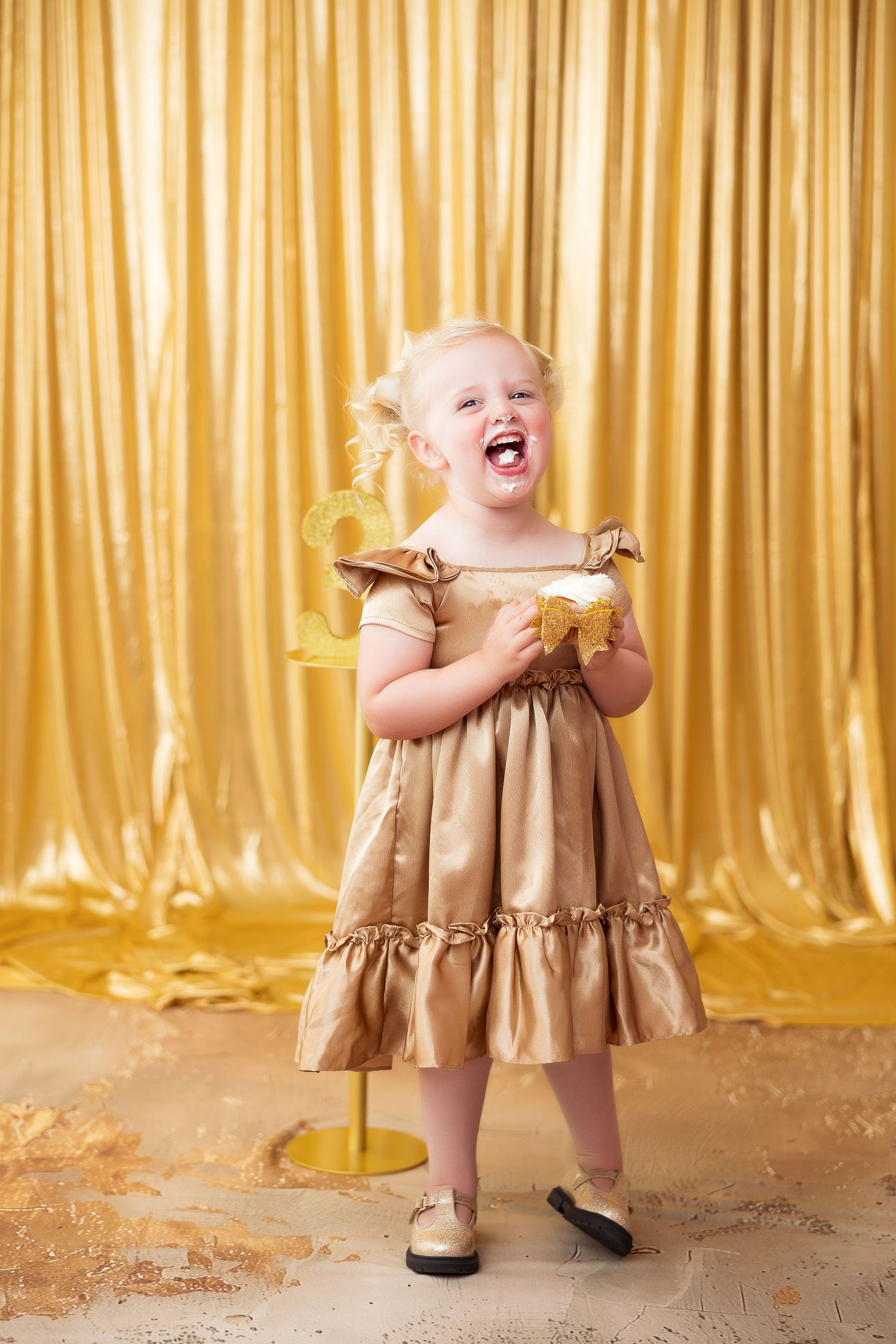 A young child wearing a gold ruffled dress laughs joyfully while standing against a shimmering gold backdrop.