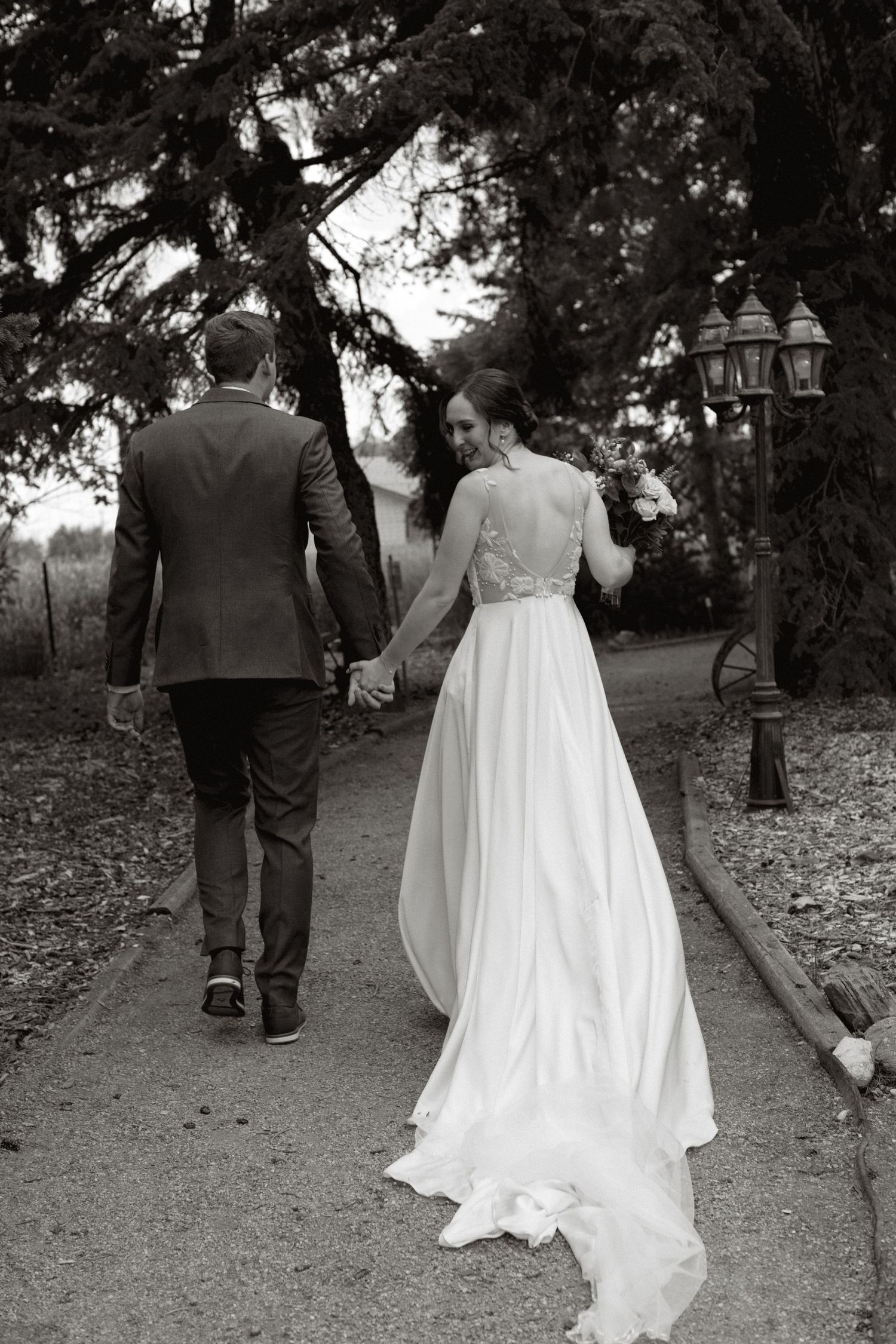 A bride and groom walk away from the camera along a tree-lined path in black and white.