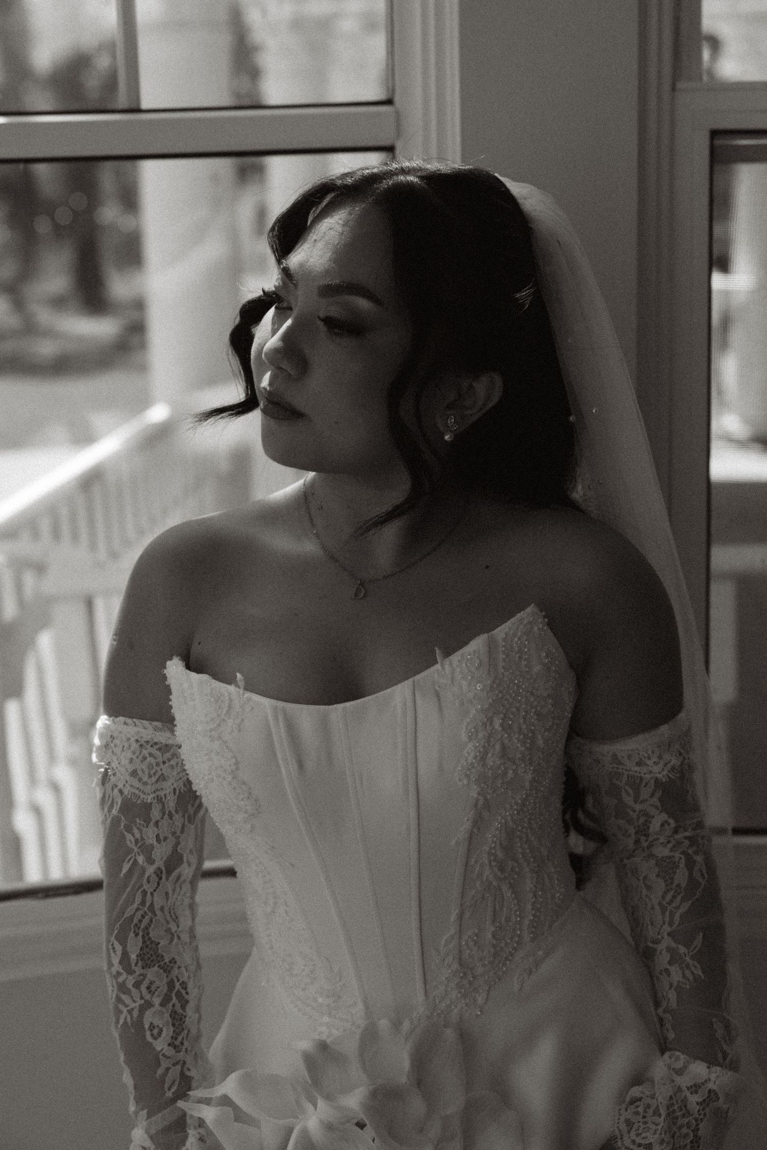 Black and white bridal portrait of a woman in a lace corset gown and veil, gazing out a window.
