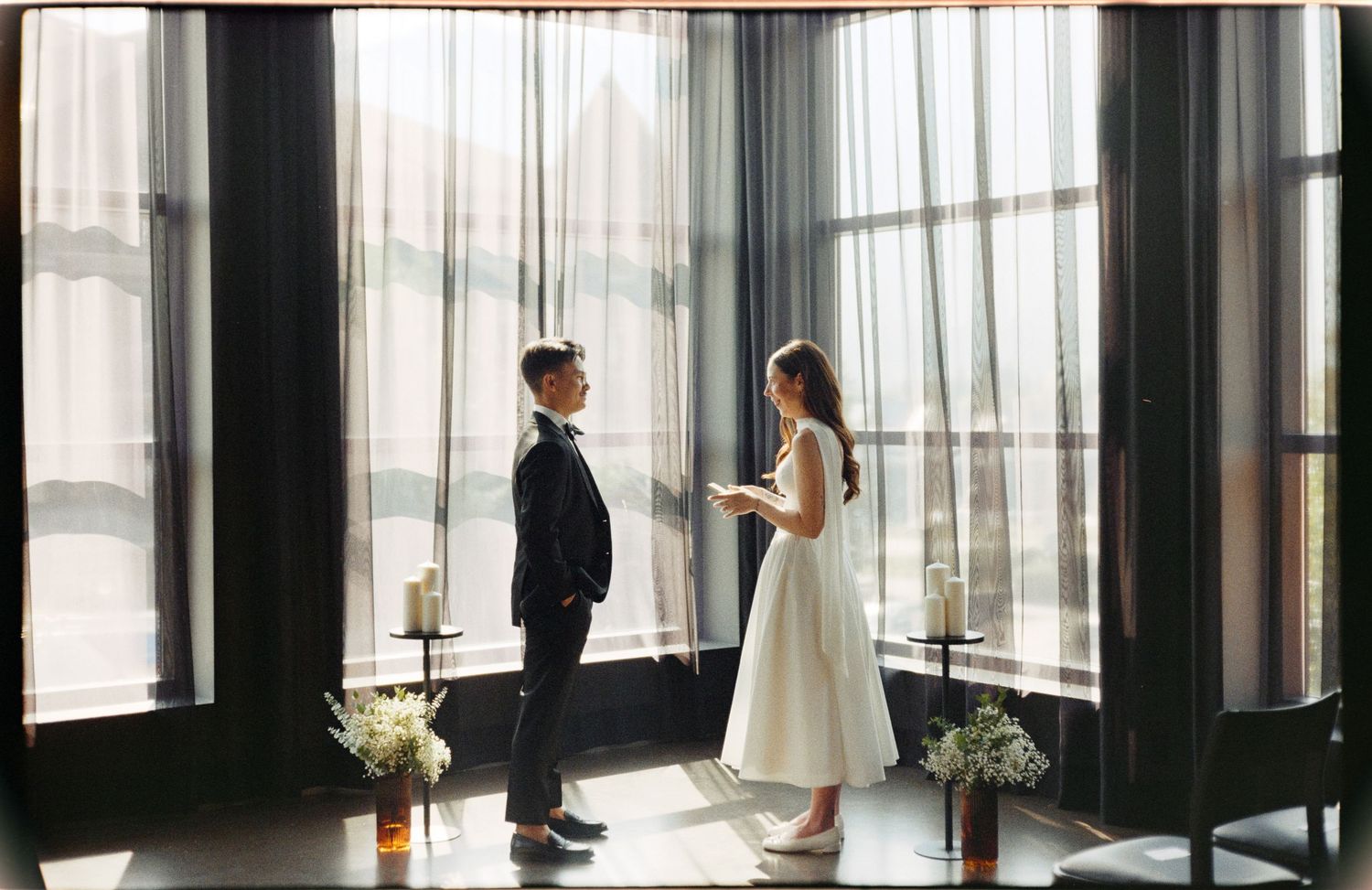 Panoramic view of couple in wedding attire by floor-to-ceiling windows.