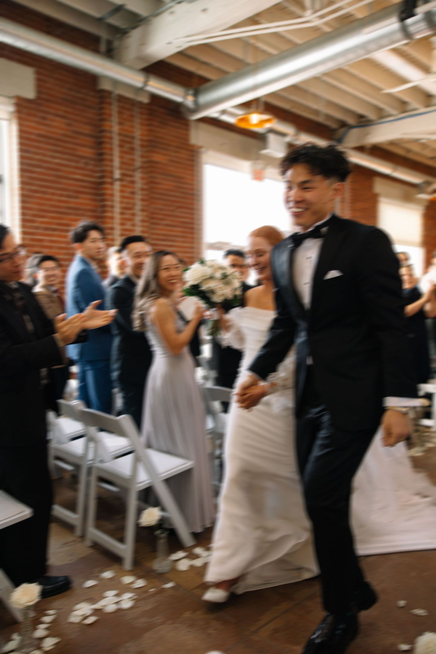 Newlyweds make their grand entrance at a rustic brick venue as guests cheer and celebrate.