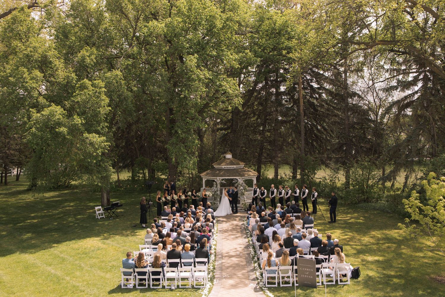 Aerial view of an outdoor wedding ceremony with white chairs arranged in rows surrounded by lush green trees.