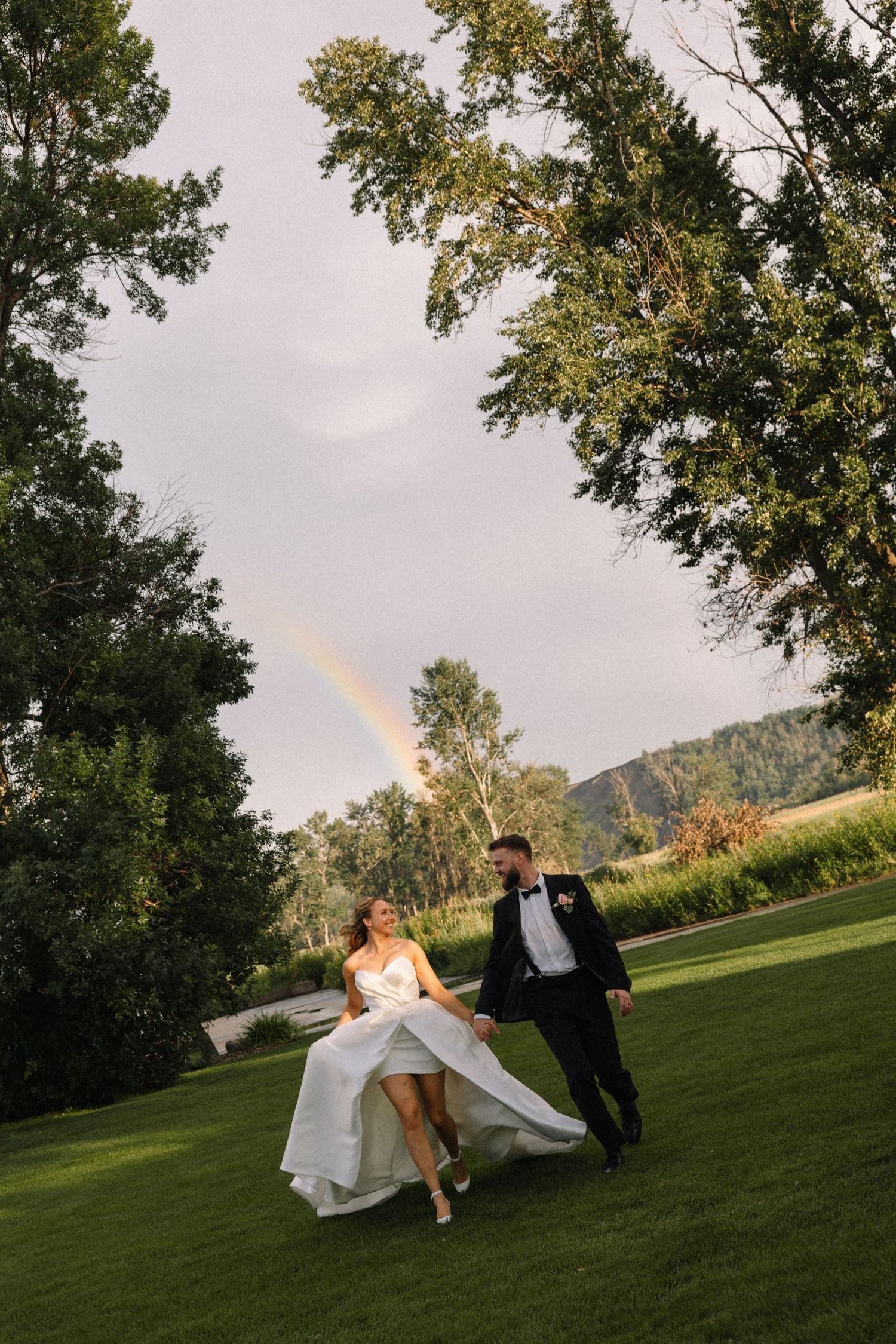 Wedding couple playfully runs across a grassy field with mountains in the background.