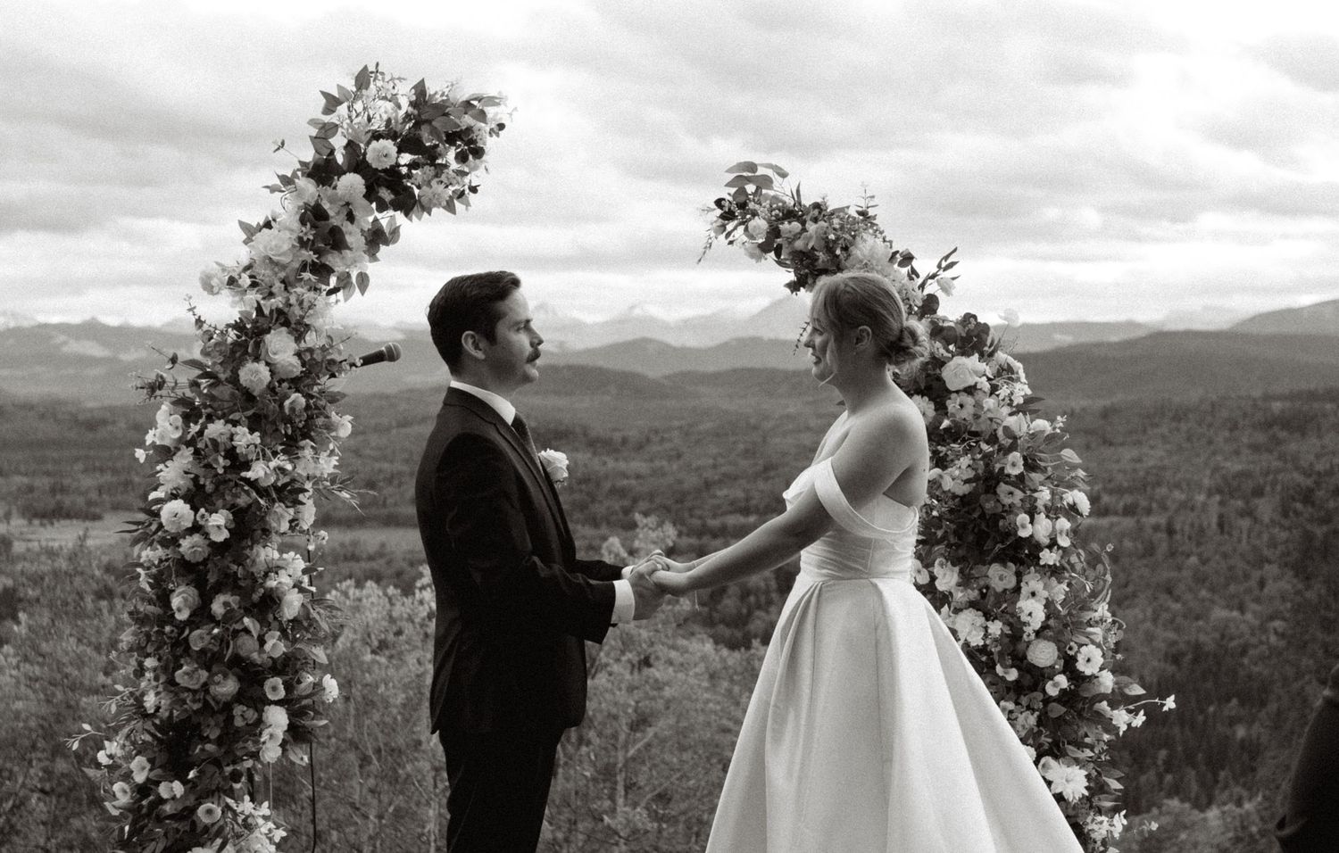 Outdoor wedding ceremony with floral arch against mountain landscape.
