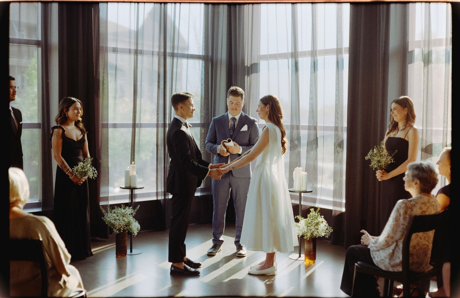 An intimate wedding ceremony takes place in a bright room with floor-to-ceiling windows and elegant black-clad attendants.