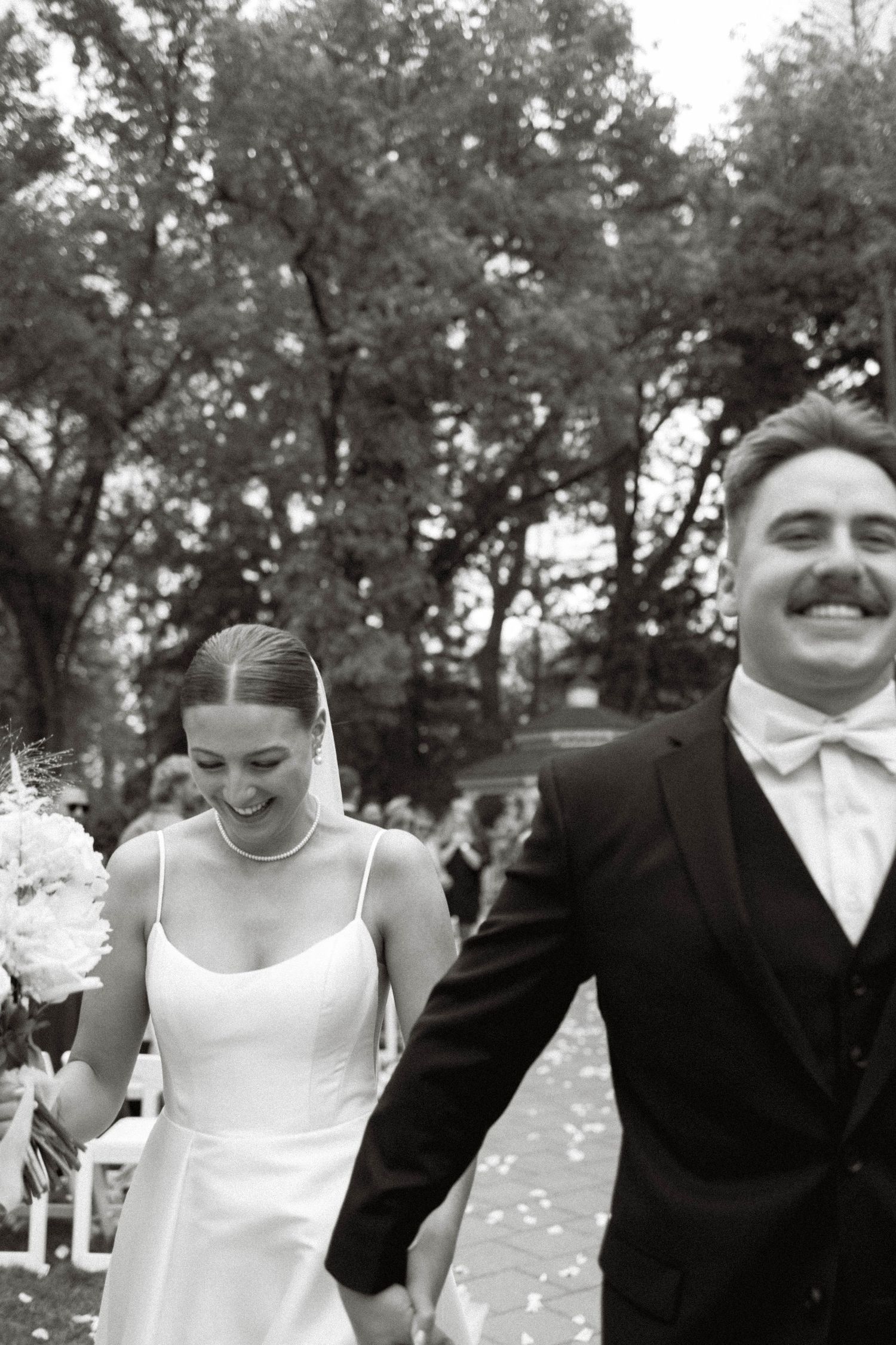 A joyful black and white photo of newlyweds walking together outdoors with trees in the background.