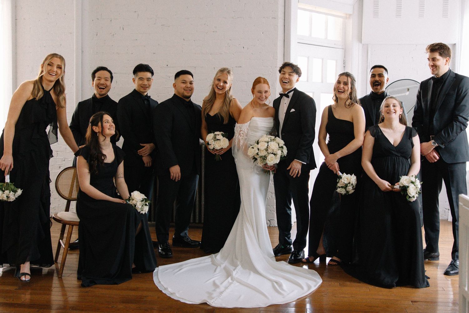 Wedding party poses in formal attire with bridesmaids in black dresses and groomsmen in dark suits.