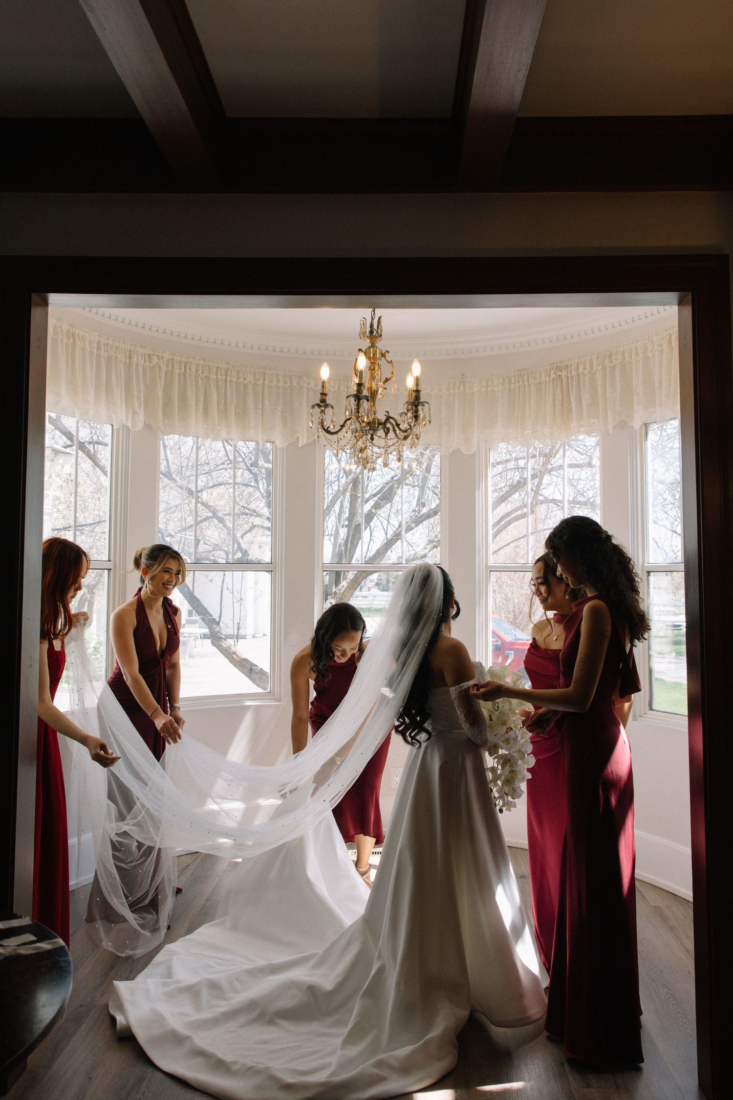 Bridesmaids in red dresses help bride with veil in elegant bay window room with chandelier.