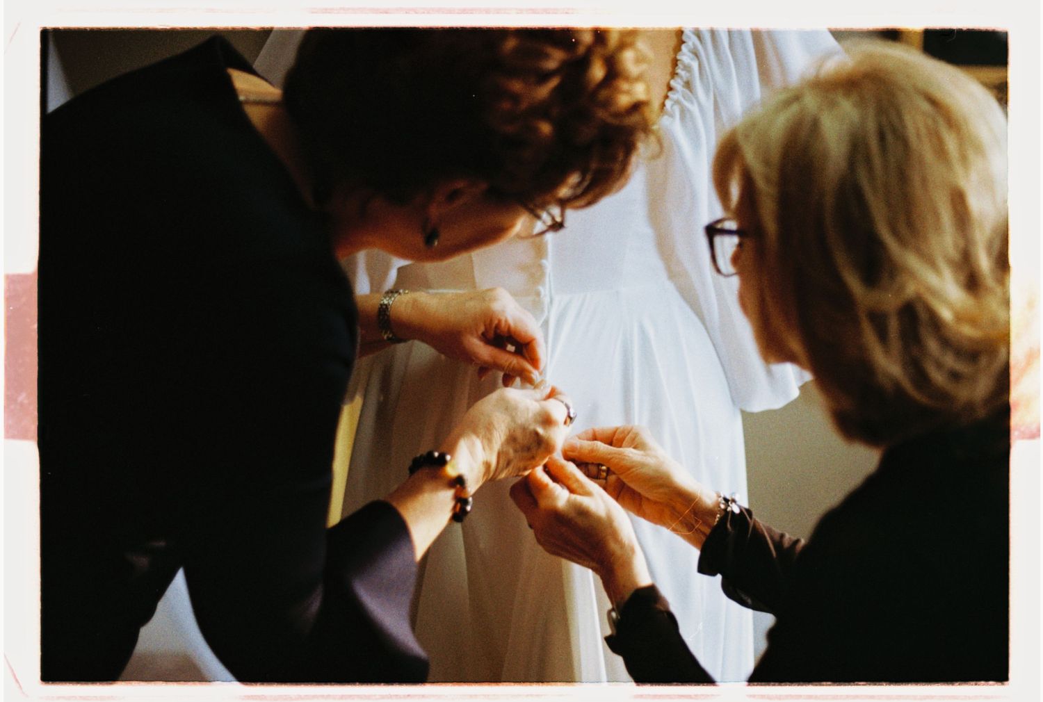 Two women helping a bride with wedding dress details during bridal preparations indoors.
