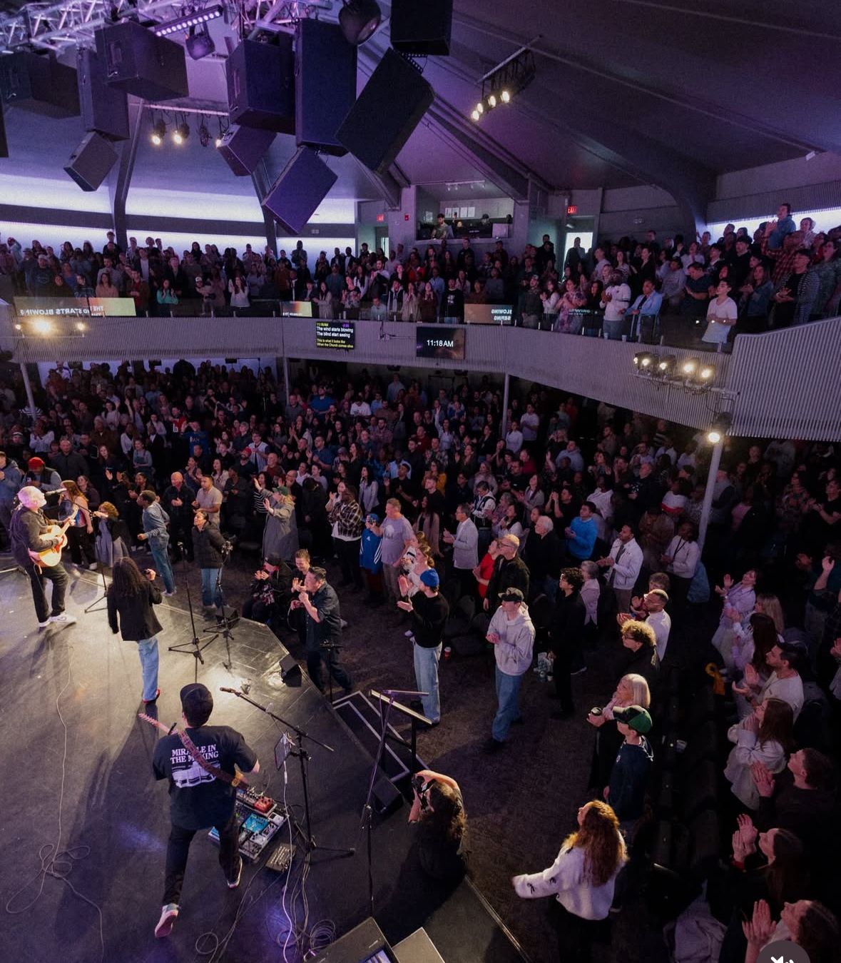 Large crowd at indoor concert venue with purple lighting and raised stage.