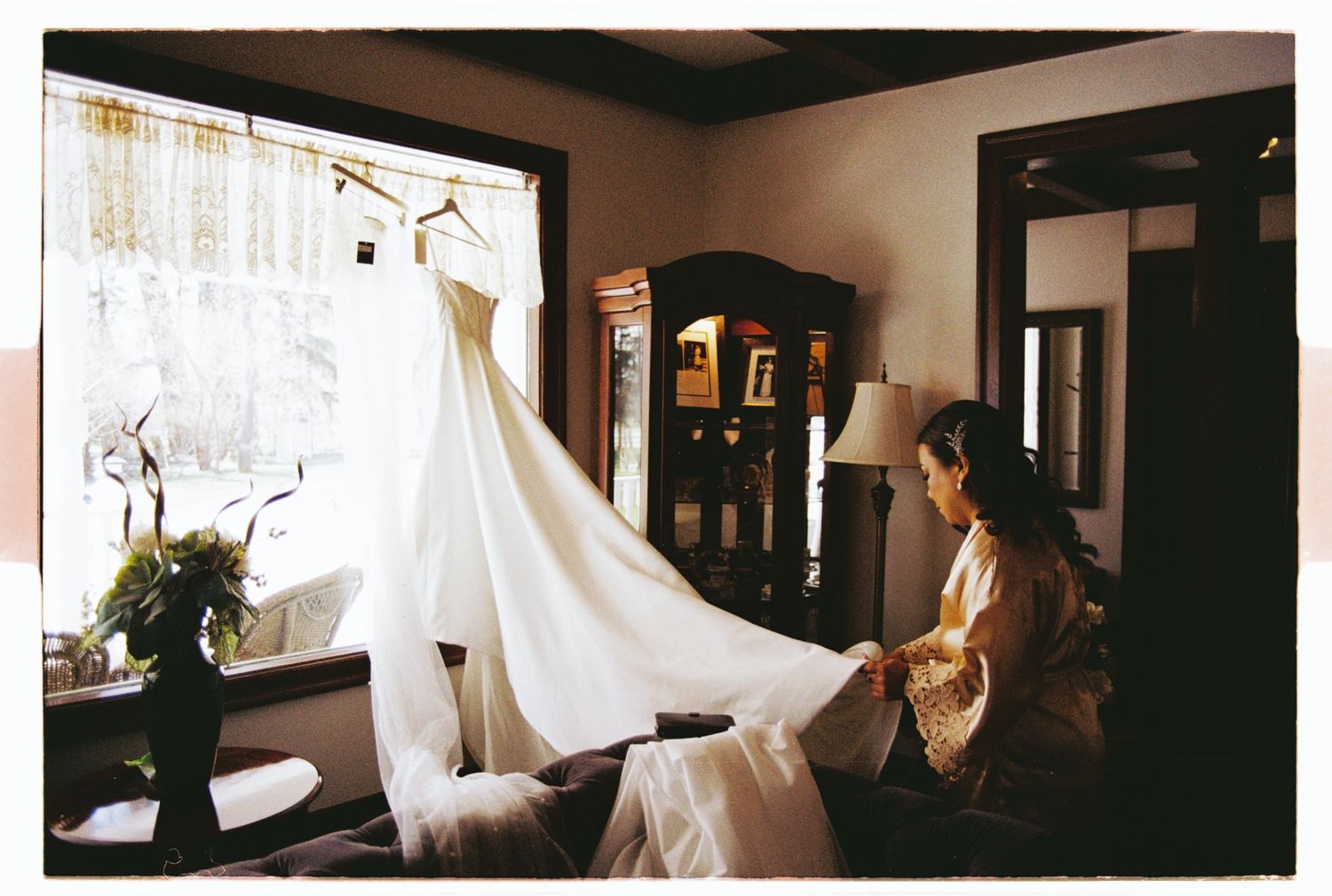 Bride sitting near window with elegant white wedding dress hanging in sunlit room before ceremony.