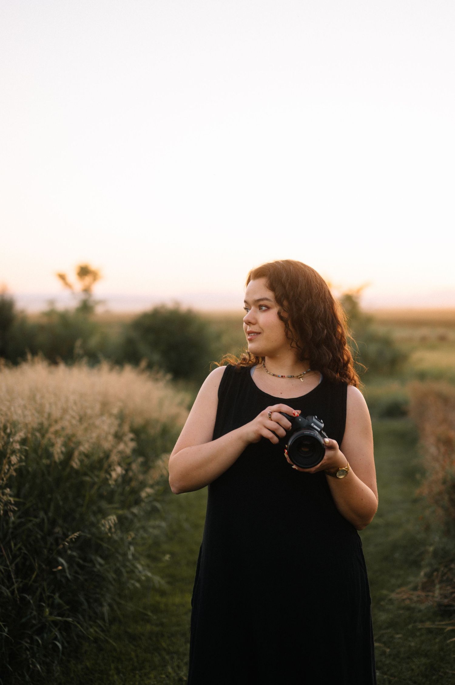 A photographer wearing black adjusts camera settings in a field at sunset.