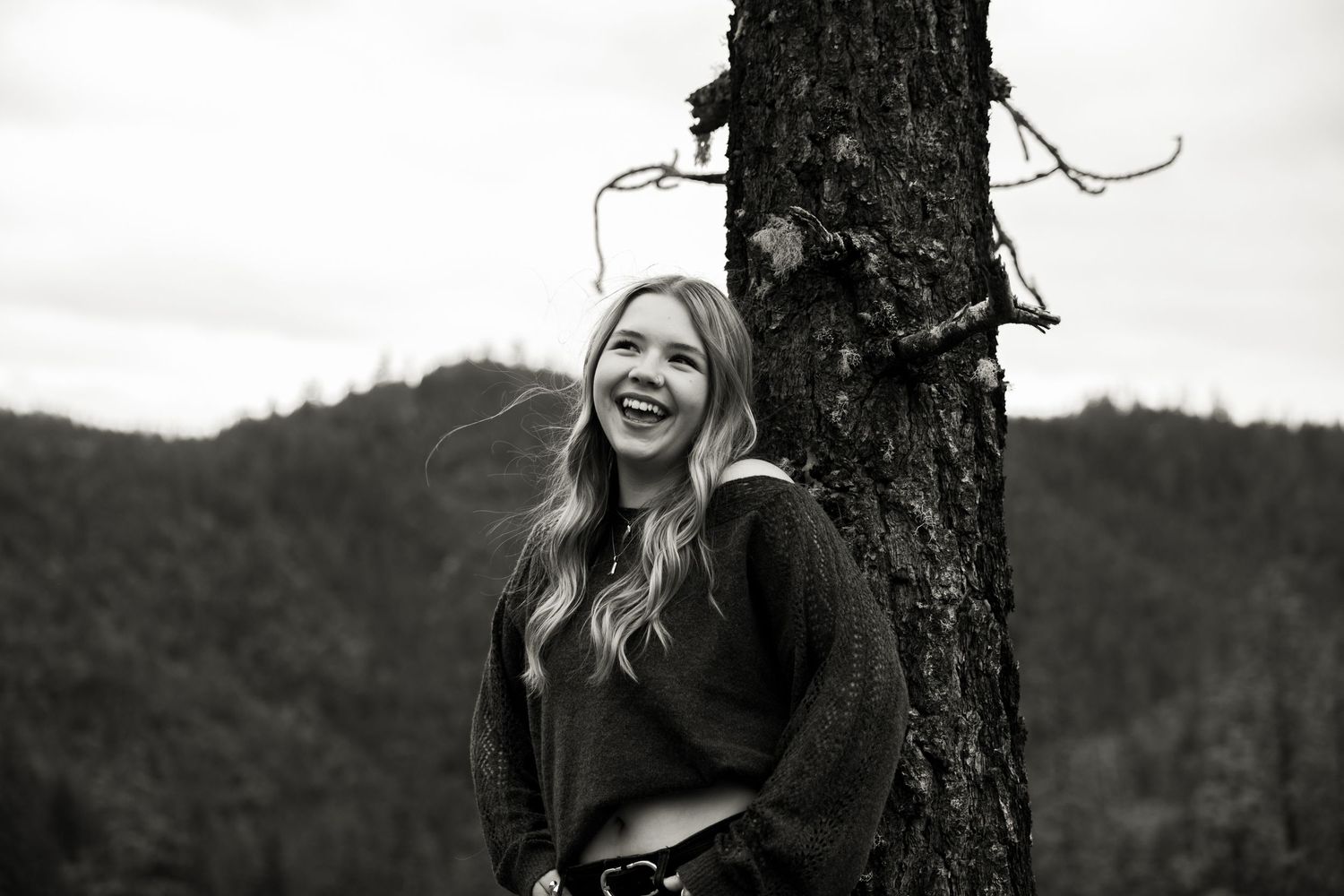 Young woman laughing beside a tree in a black and white outdoor portrait with forested hills behind.