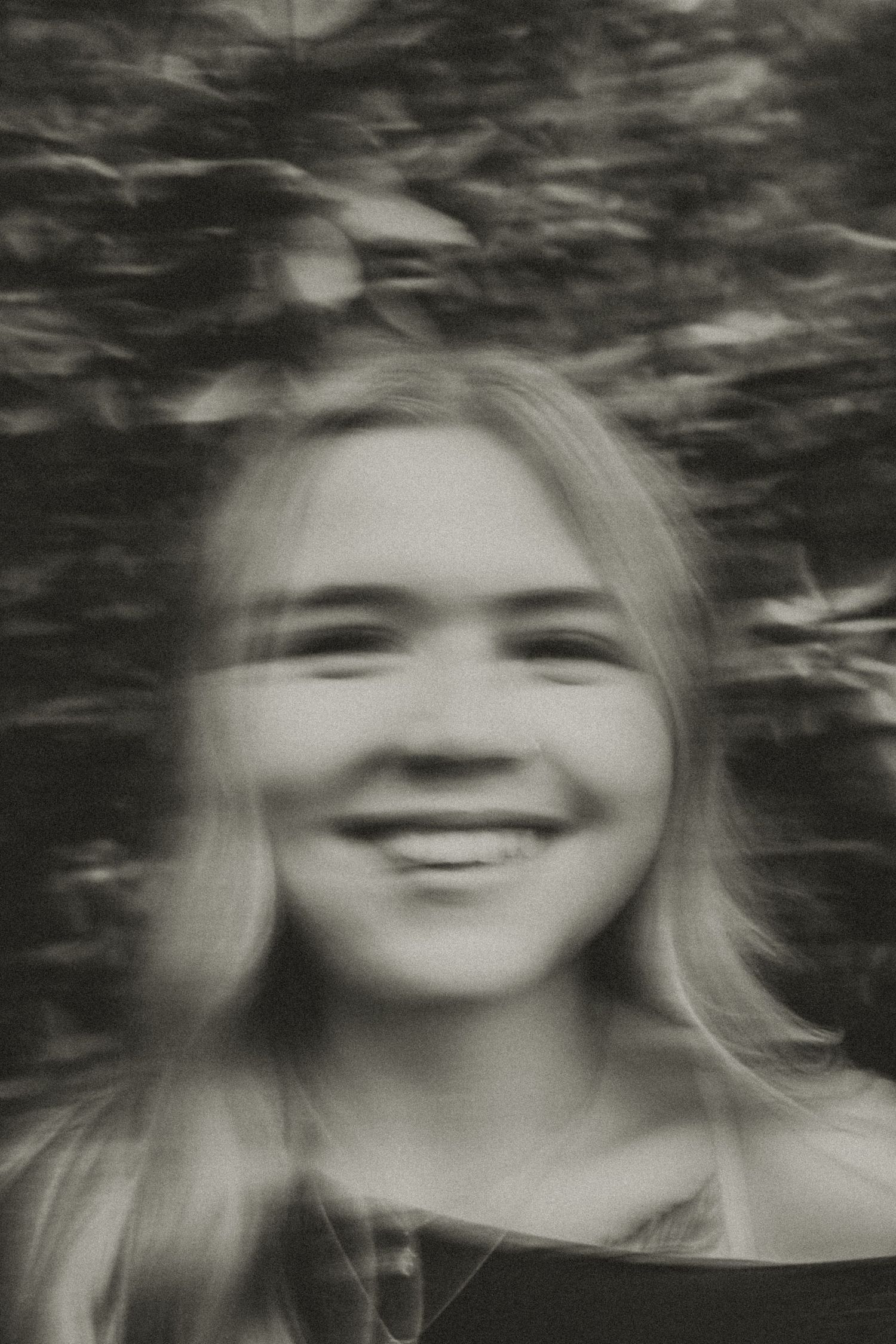 Blurry black and white portrait of a smiling young blonde woman outdoors with trees in background.