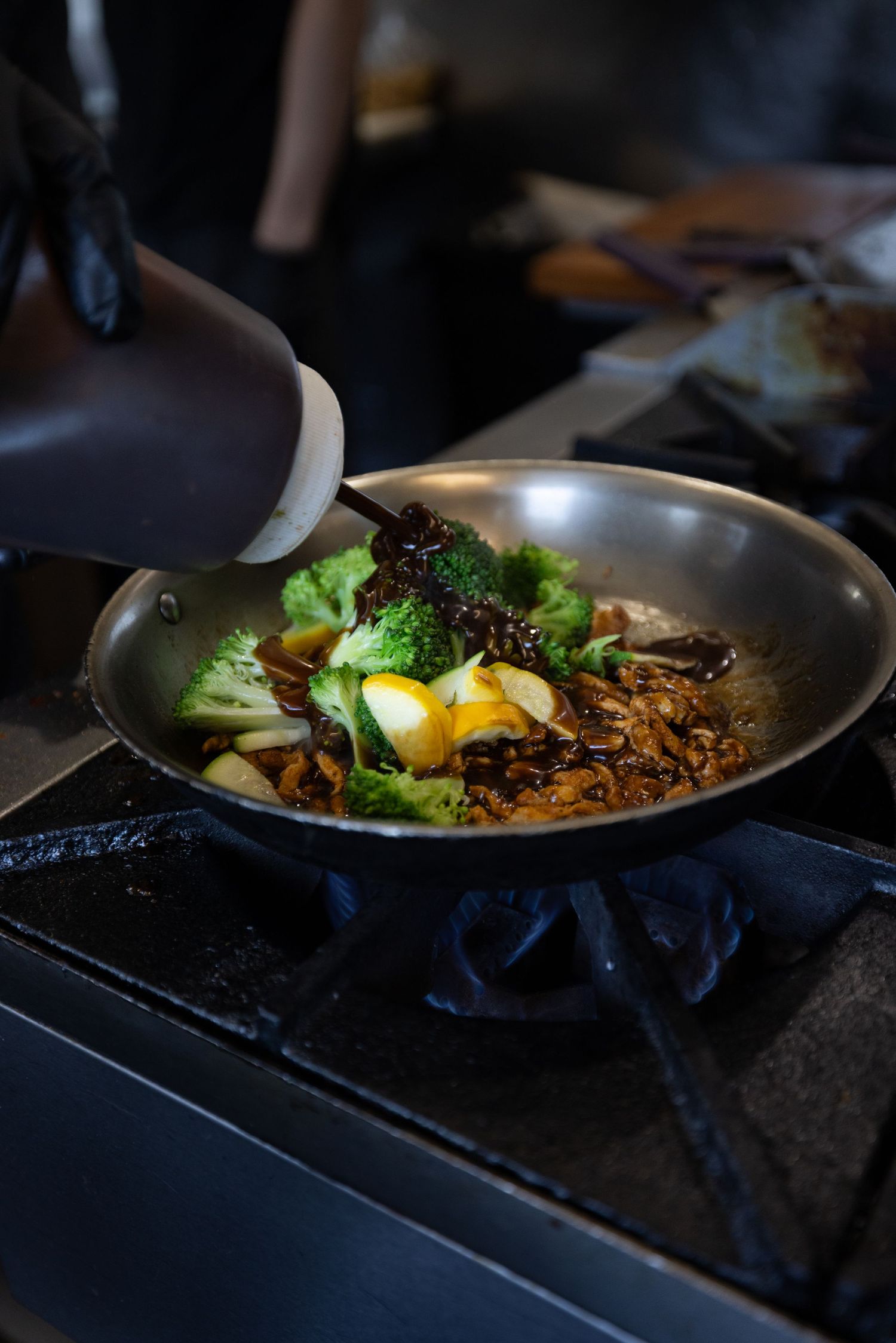 Chef adding sauce to a stir-fry with broccoli, eggs, and mushrooms in a steel wok on a commercial stove.