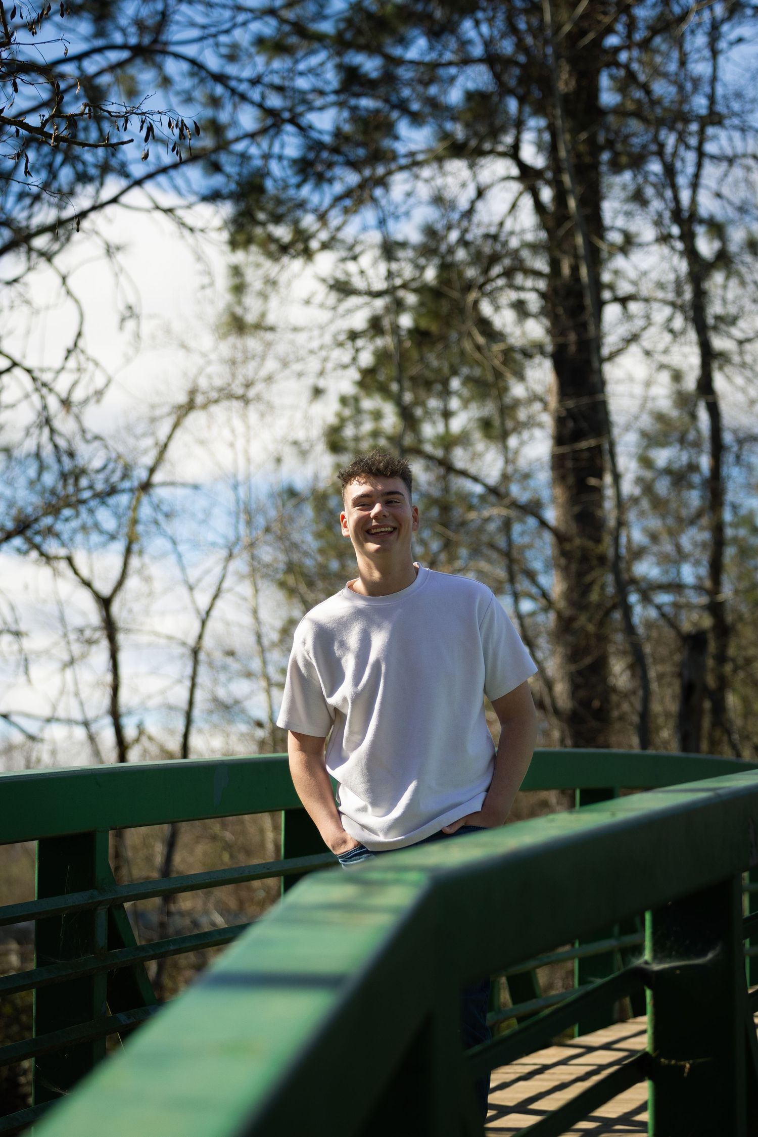 Young man in white t-shirt smiling on a green bridge surrounded by trees on a sunny day.