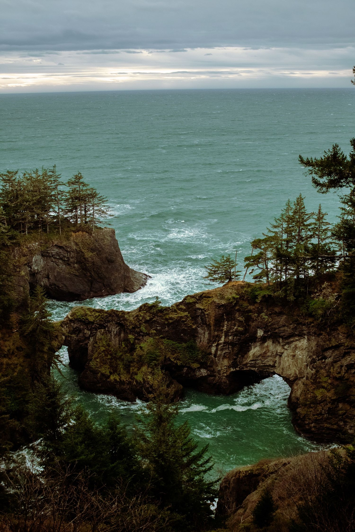 Rocky coastal arch over turquoise waters with evergreen trees on cliffs under cloudy skies on the Oregon coast.
