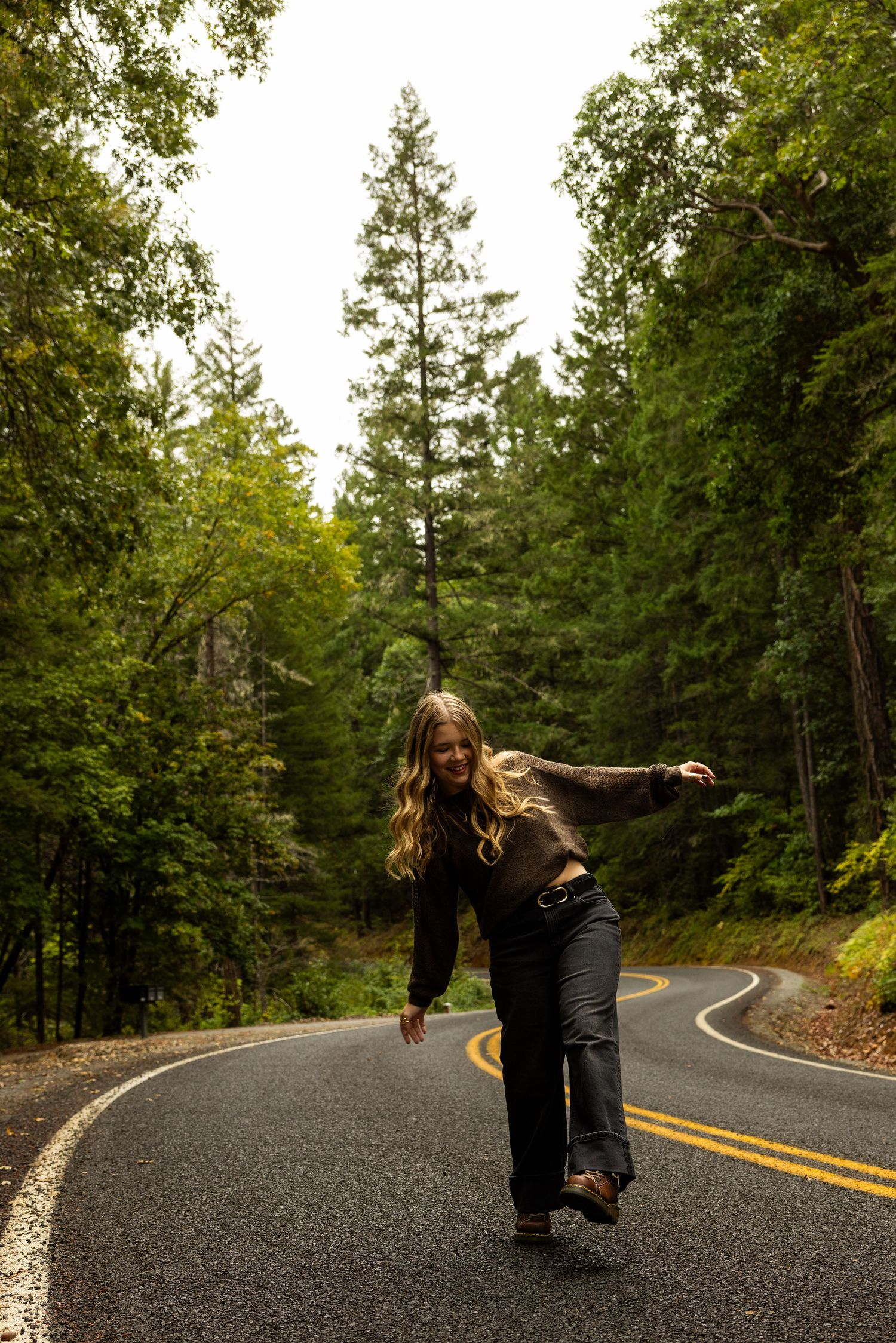 Woman balancing on winding forest road surrounded by tall green trees, wearing dark jeans and brown sweater.