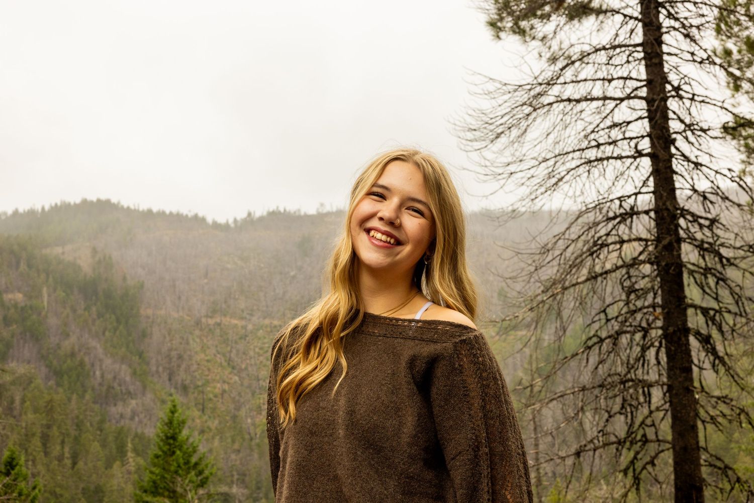 Smiling young woman with long blonde hair wearing a brown sweater, standing outdoors with misty forested mountains behind her.