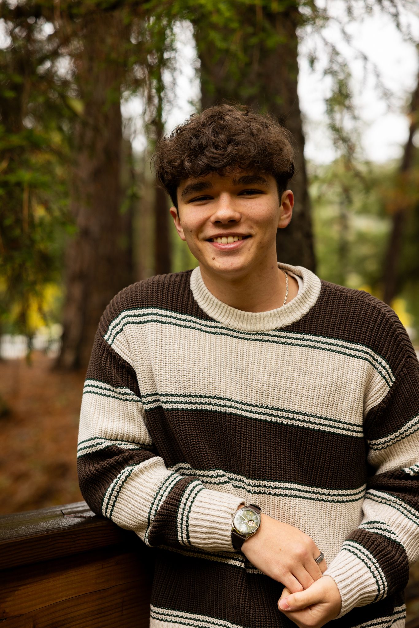 Young man in striped brown sweater smiling outdoors in a forest setting, wearing a watch.