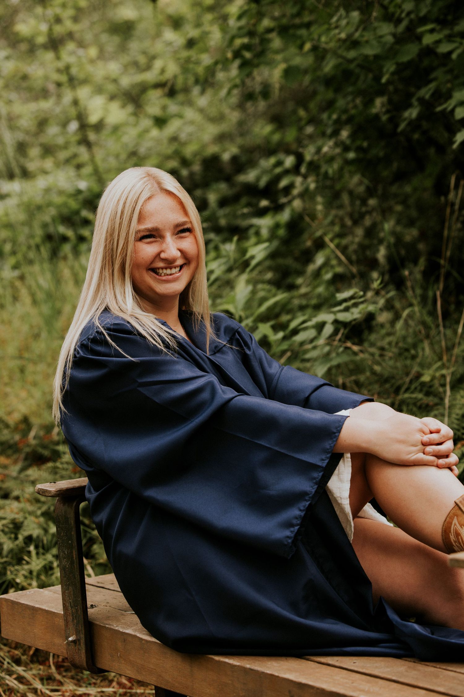 A person in a navy graduation gown relaxes on a wooden bench surrounded by greenery in an outdoor setting.