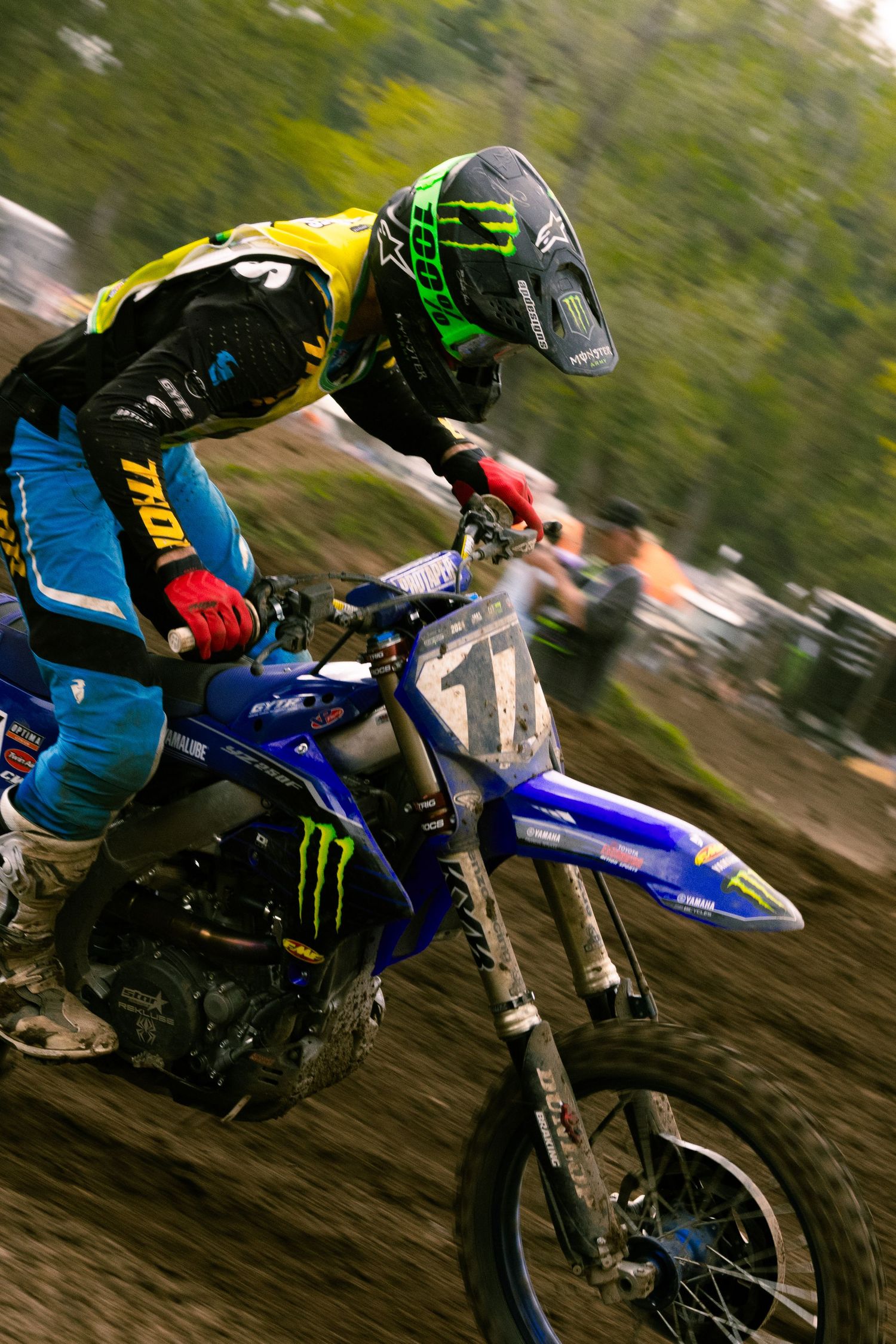 Motocross rider in Monster Energy gear racing a blue Yamaha dirt bike on a muddy track during competition.