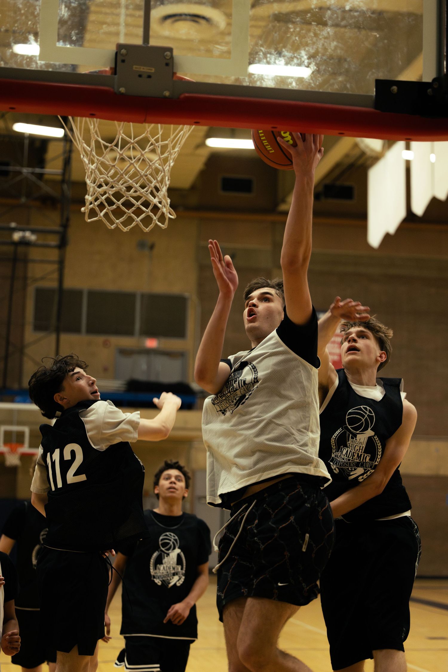 A young basketball player in a white jersey drives to the hoop between two defenders in black uniforms during an indoor game.