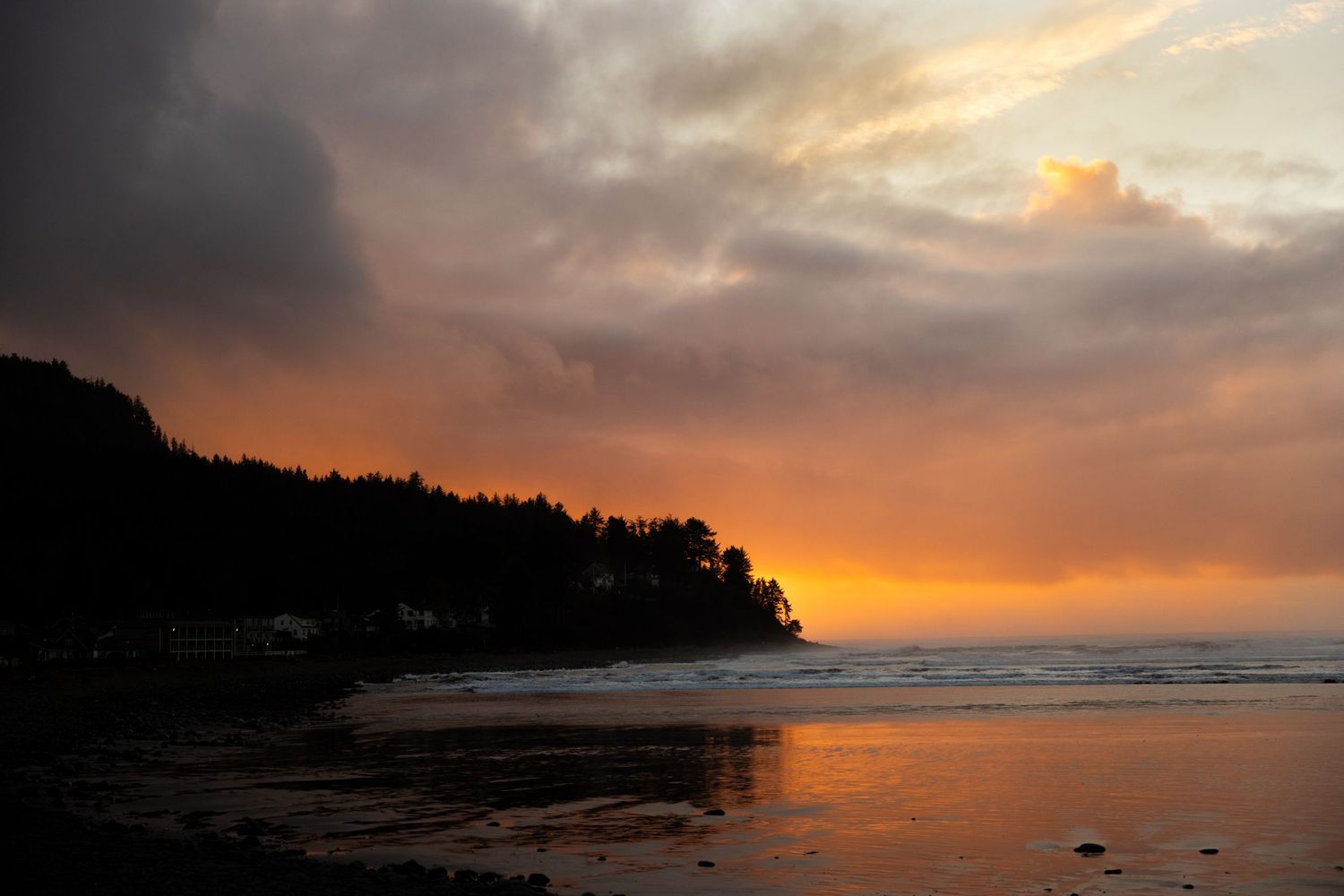 Dramatic sunset over a coastal headland with glowing orange sky reflecting on calm ocean waters and silhouetted evergreen trees.