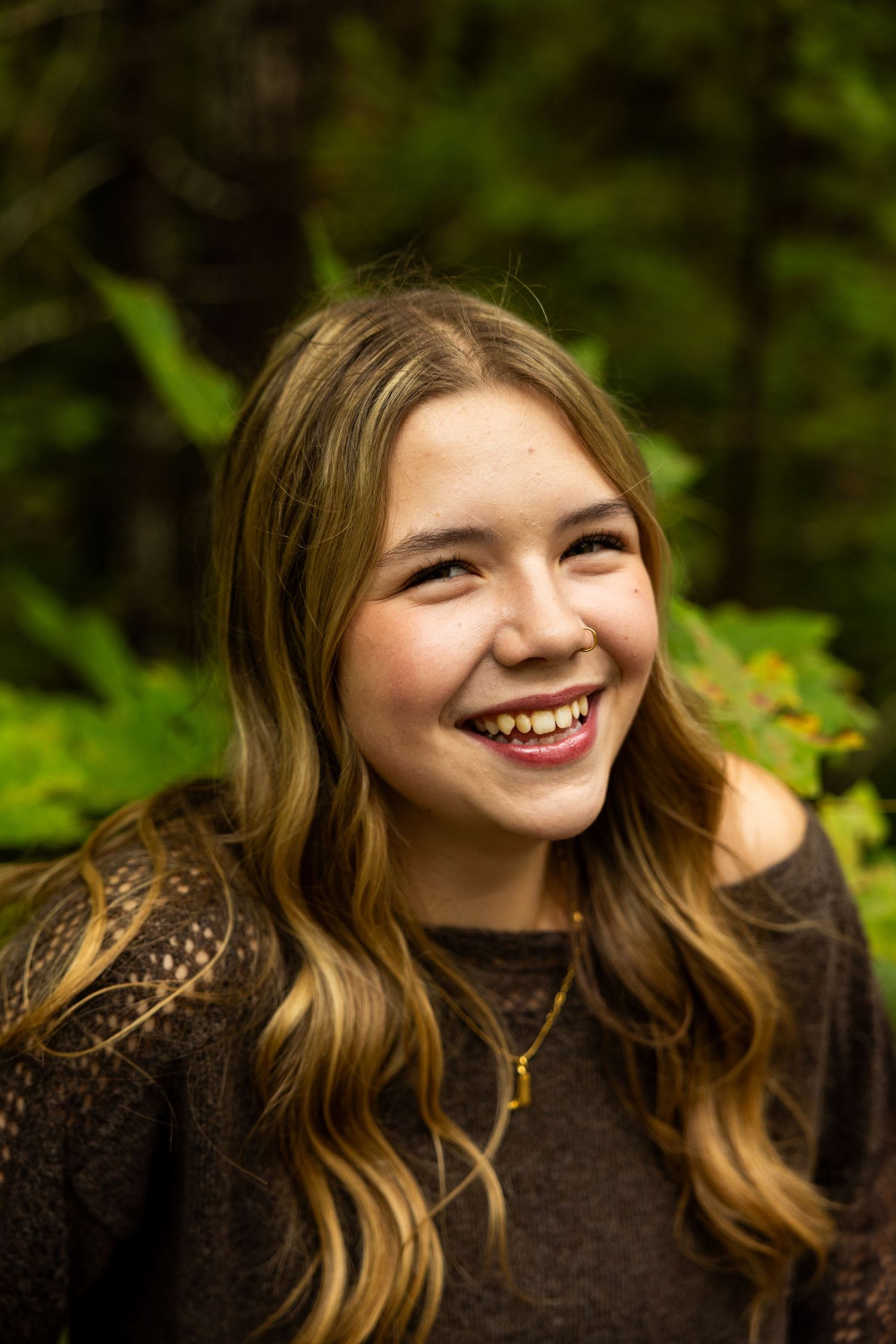 Smiling teenage girl with long wavy blonde hair wearing a dark top, laughing outdoors with green foliage background.