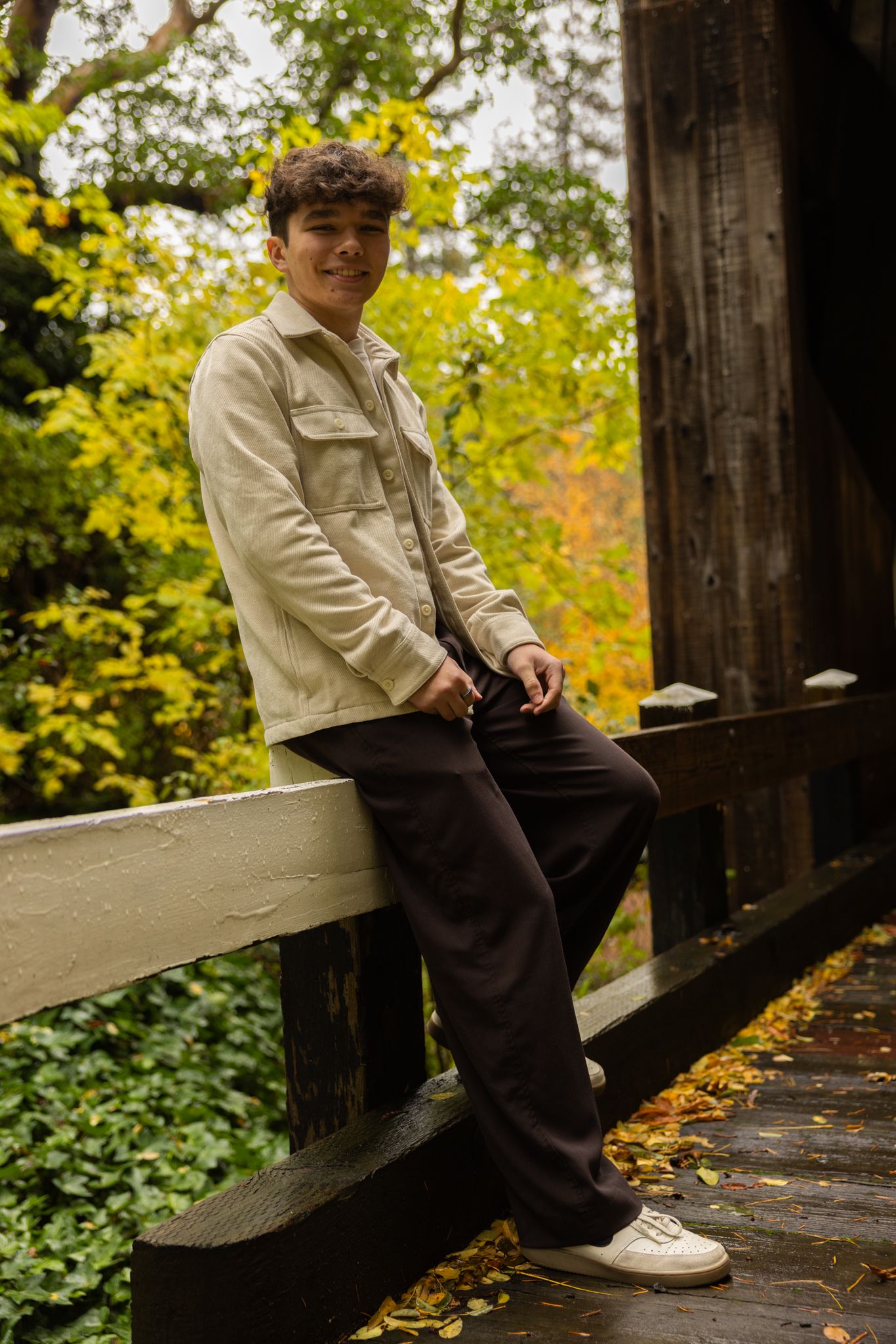 Young man in beige jacket and dark pants leaning on wooden bridge railing surrounded by autumn foliage.