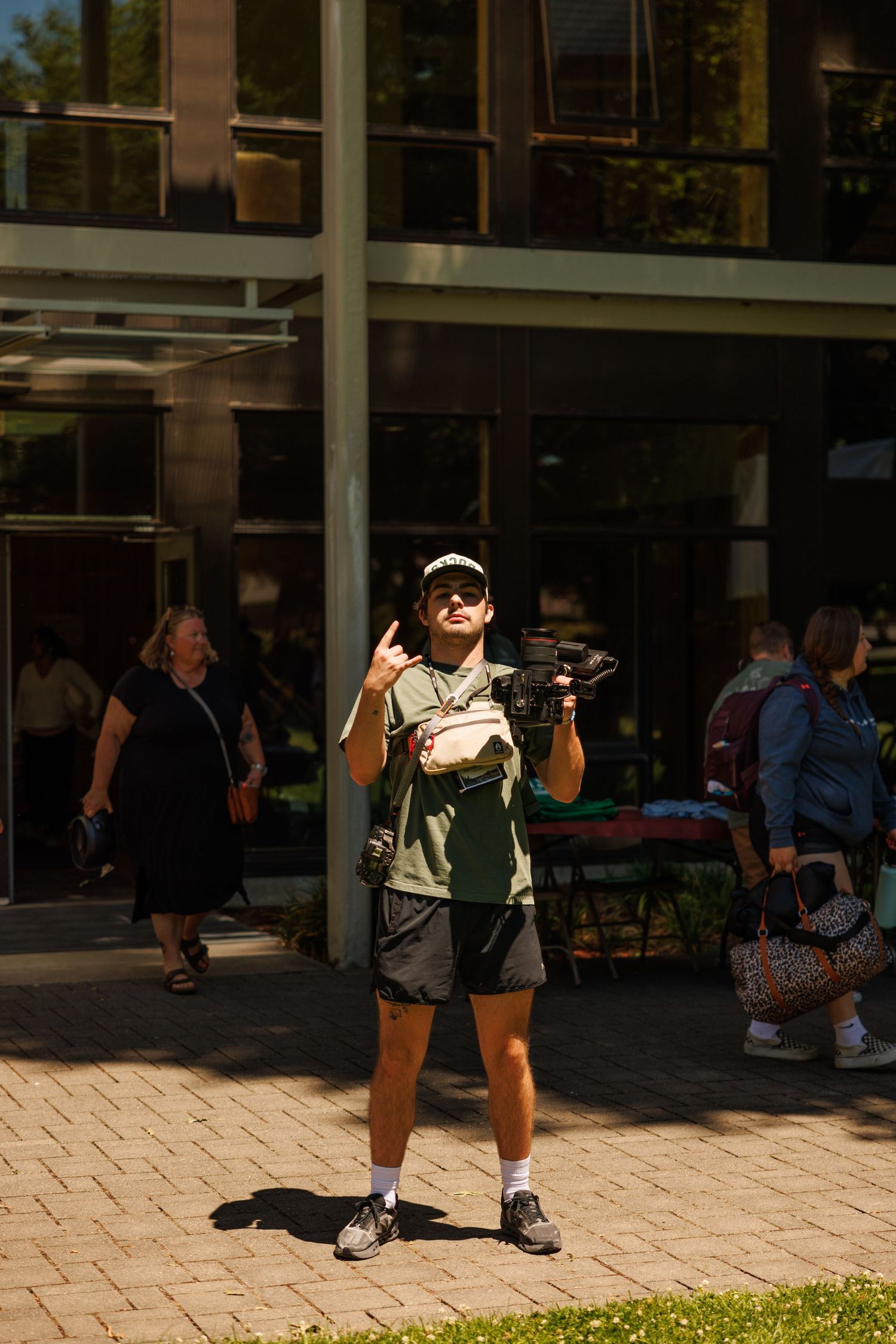 Young man in green shirt and cap holds camera outdoors, pointing up near a modern glass building on a sunny day.