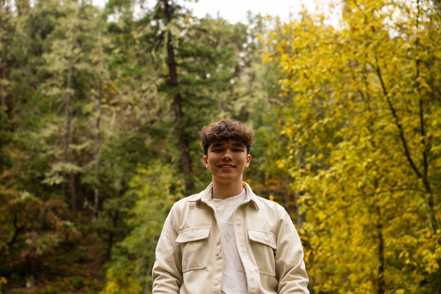 Young man smiling in a forest with vibrant yellow and green autumn trees in the background.