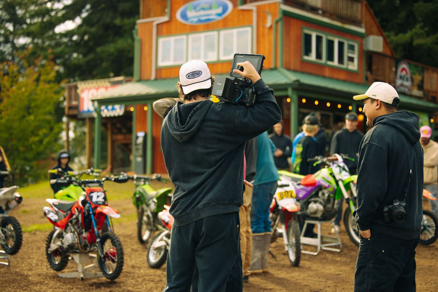 A row of dirt bikes parked at a motorsports event with orange buildings and spectators in the background.