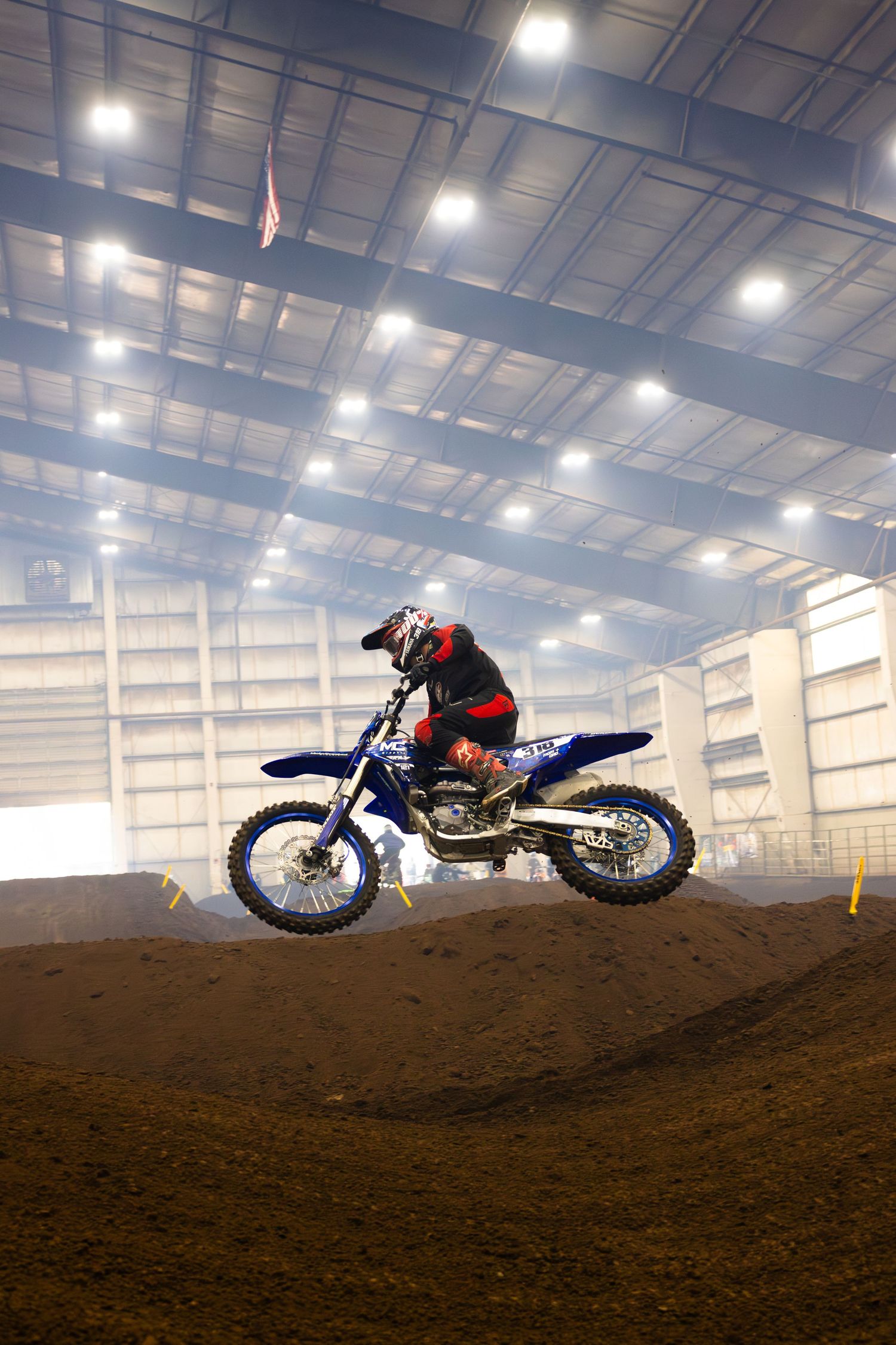 Motocross rider on a blue dirt bike catches air over an indoor supercross track under bright arena lights.