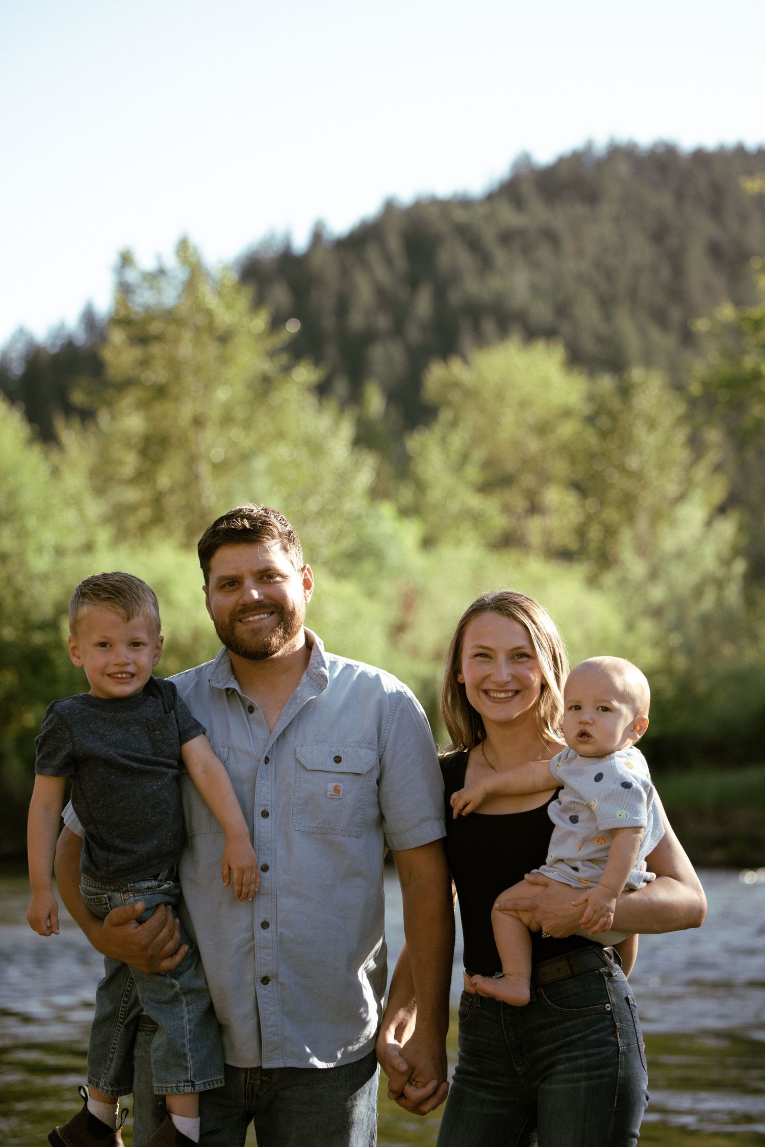 A family stands together smiling along a riverbank with trees and forest in the background.