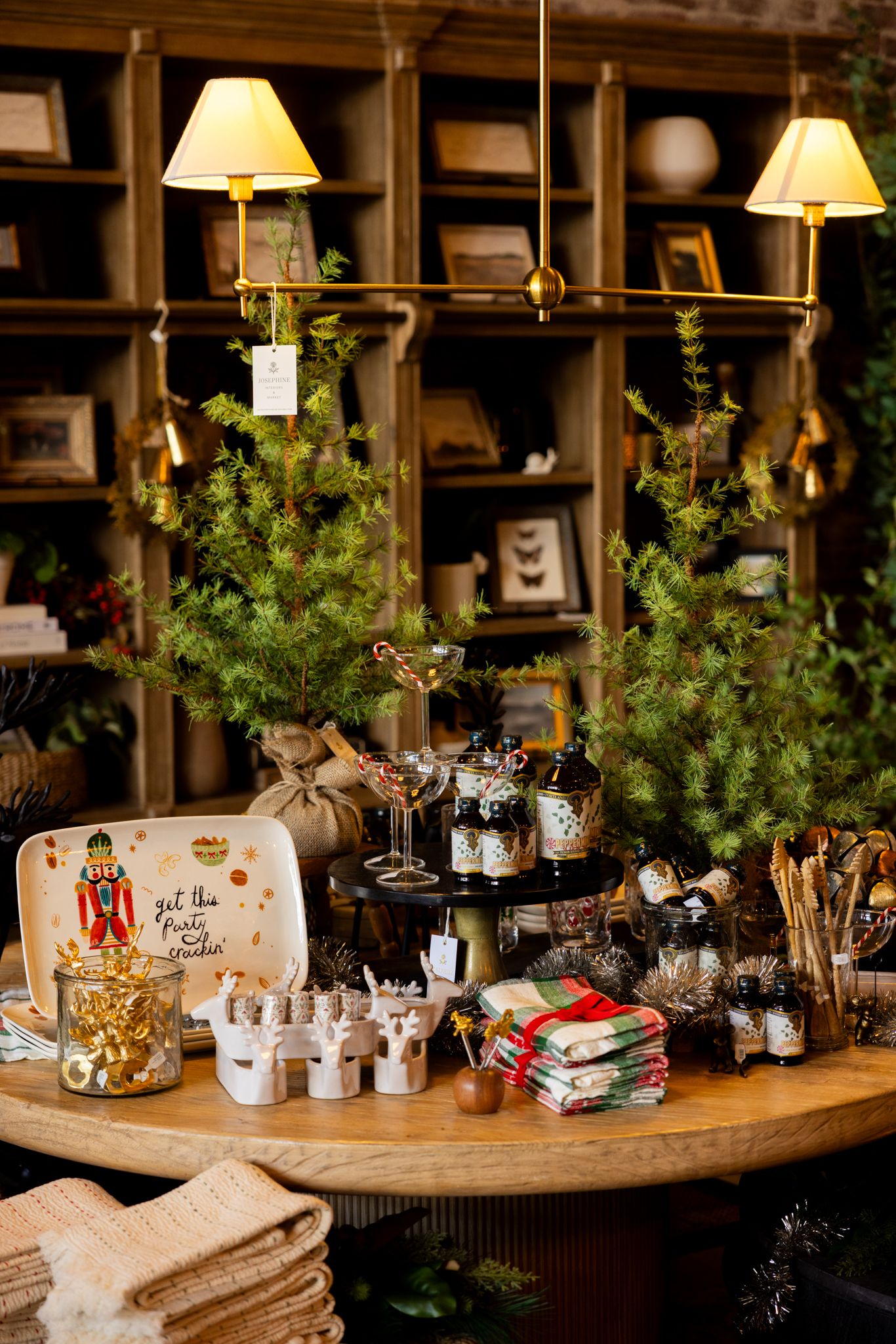Cozy holiday display with small Christmas trees, decorative items, and table settings arranged on wooden surface with bookshelves.