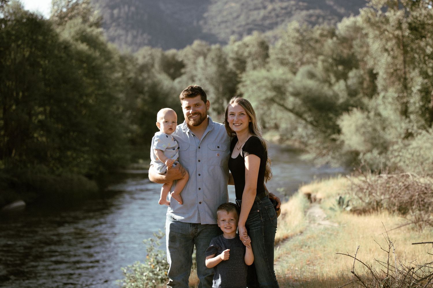 Family poses together on riverbank with mountains and trees in background.