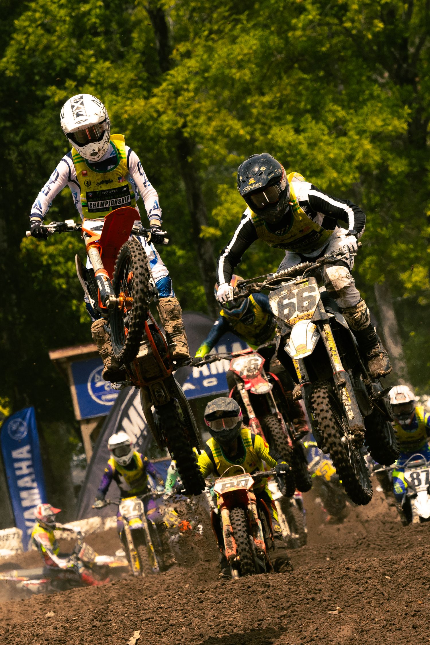 Motocross riders competing on dirt track with number 66 bike leading the race, green trees in background.