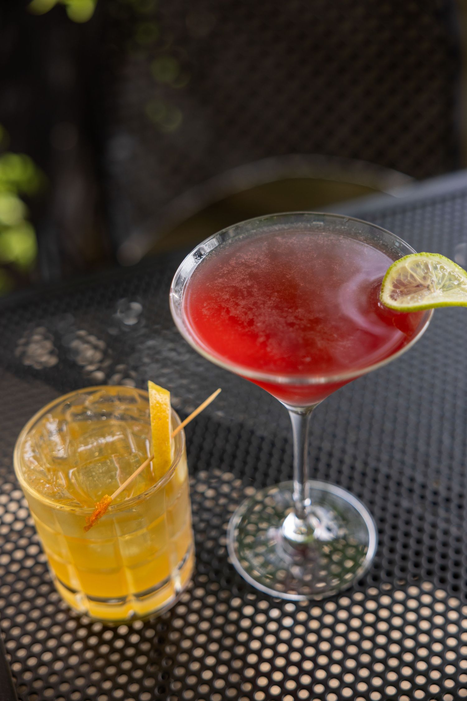 Two cocktails on a dark mesh table, a red martini with lime and a yellow whiskey sour with lemon peel.