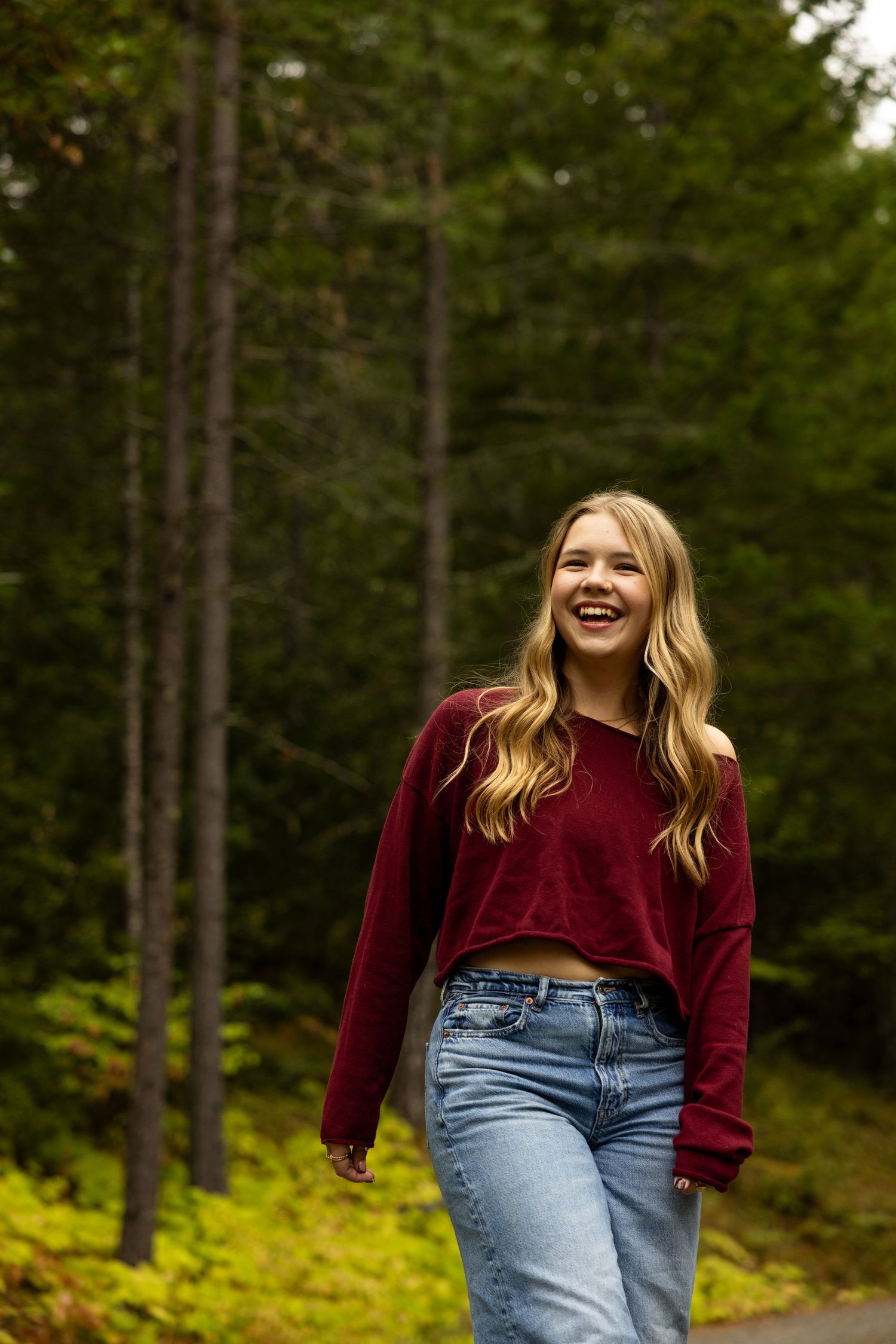 Smiling young woman in red crop top and jeans walking on a forest path surrounded by tall green trees.