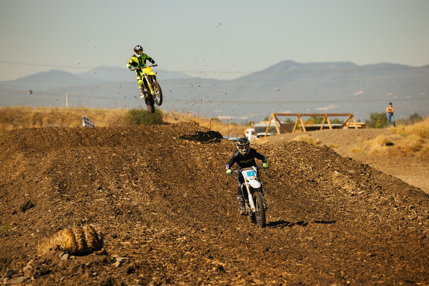 Motocross riders jumping over dirt mounds on a dusty track with mountains visible in the background.